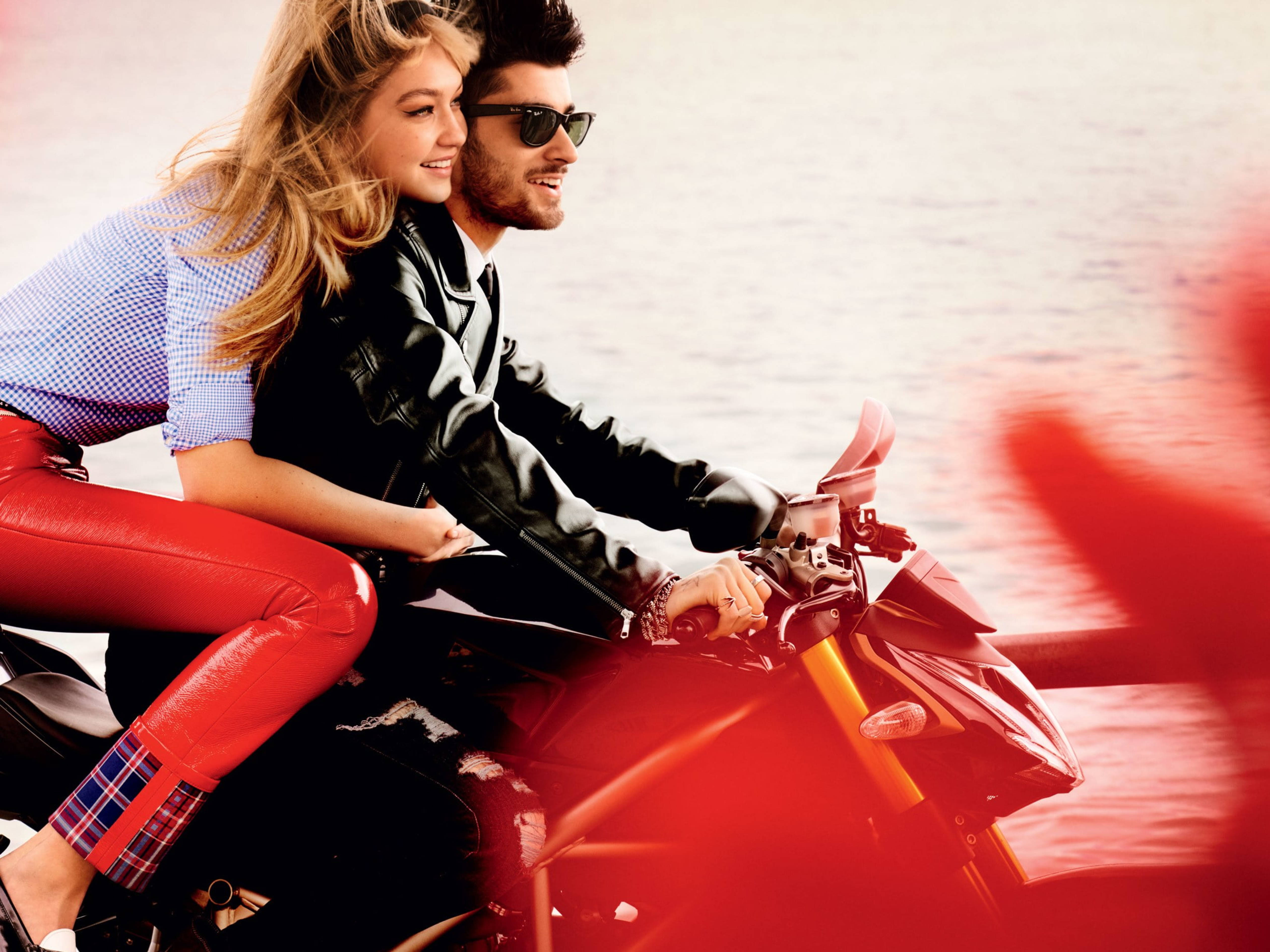 zayn 桌布 壁紙 — 一對微笑的情侶在黃金時段騎著紅色機車，沿著模糊的海濱行駛，頭髮隨風飄揚。