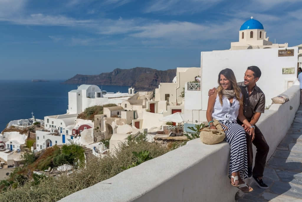 Greek Island 桌布 壁紙 — 一對微笑的情侶坐在白色石牆上，俯瞰著伊亞聖托里尼標誌性的藍頂教堂和白色建築，背景是明亮的藍天。