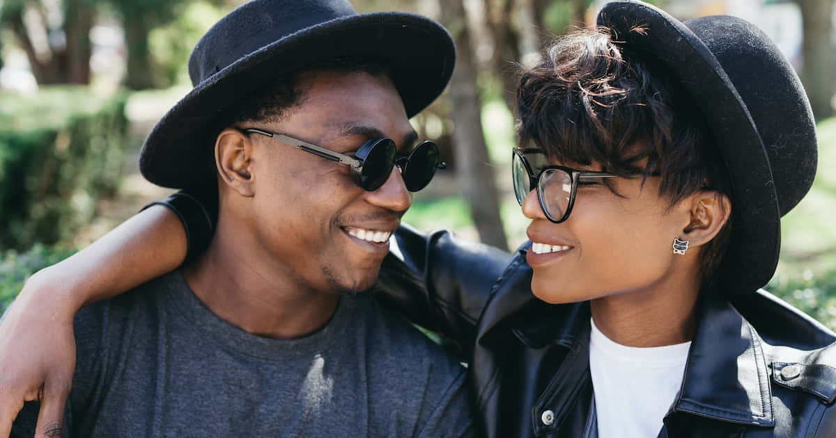 Black Couple 桌布 壁紙 — 一對微笑的情侶戴著黑色帽子和太陽眼鏡，在陽光普照的公園裡溫暖擁抱，身後有樹木。