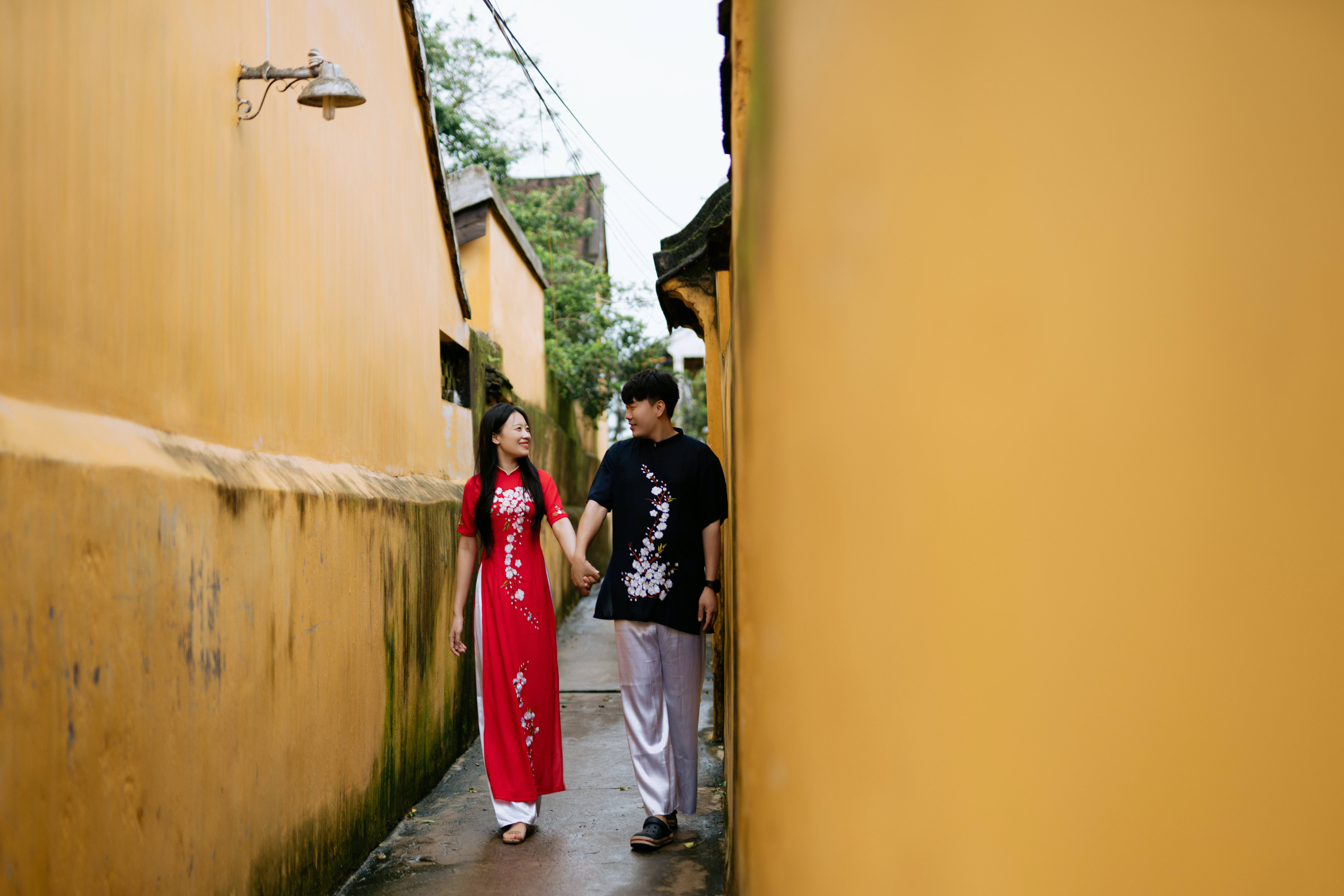 Couple 桌布 壁紙 — 一對微笑的情侶手牽手走在一條狹窄的小巷中，兩旁是溫暖的黃色牆壁，沐浴在柔和的白日光線下。