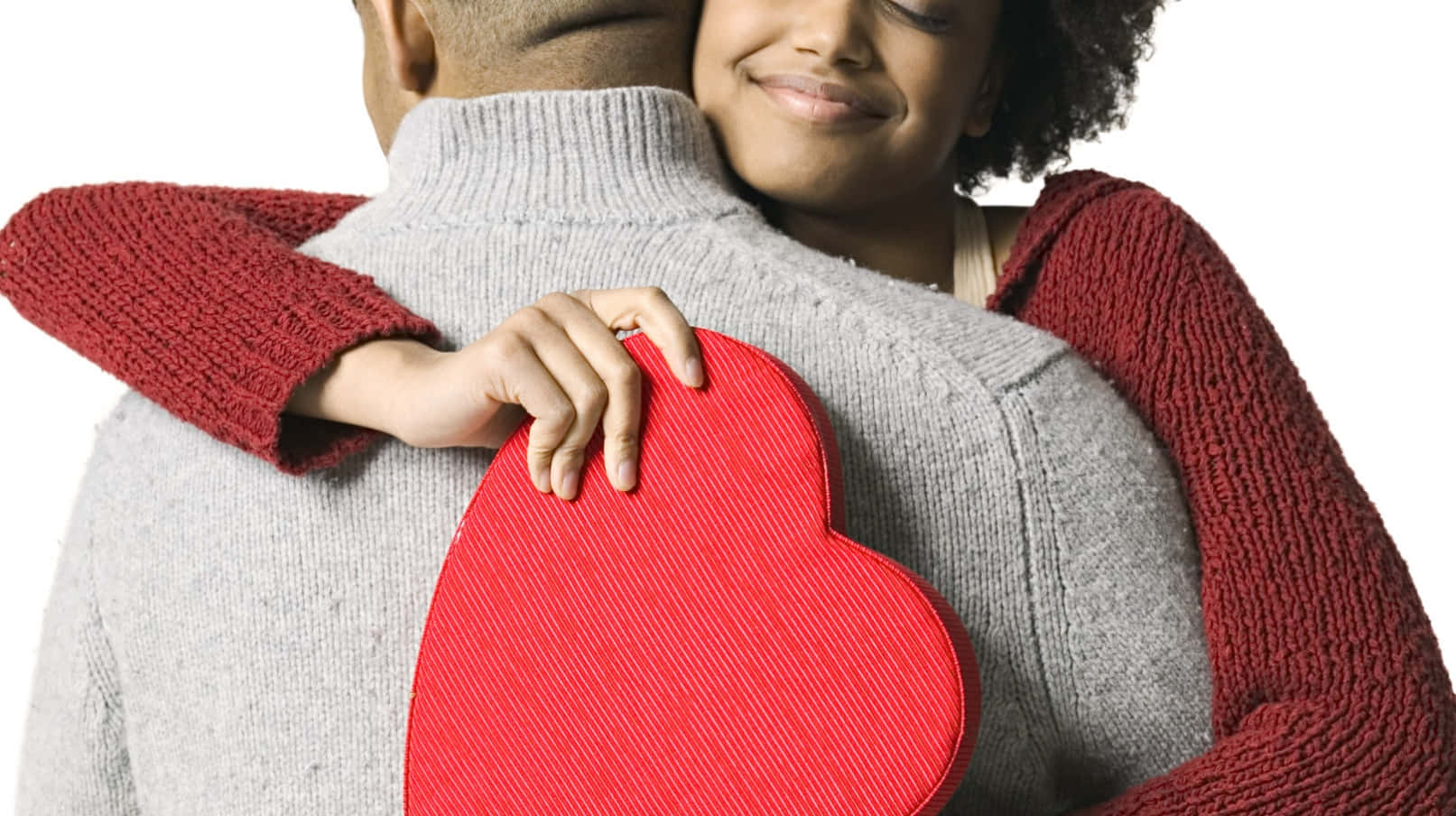 Valentine's Day 桌布 壁紙 — 一對微笑的情侶溫暖地擁抱，女士拿著一個紅色心形盒子靠在男士的灰色毛衣上。