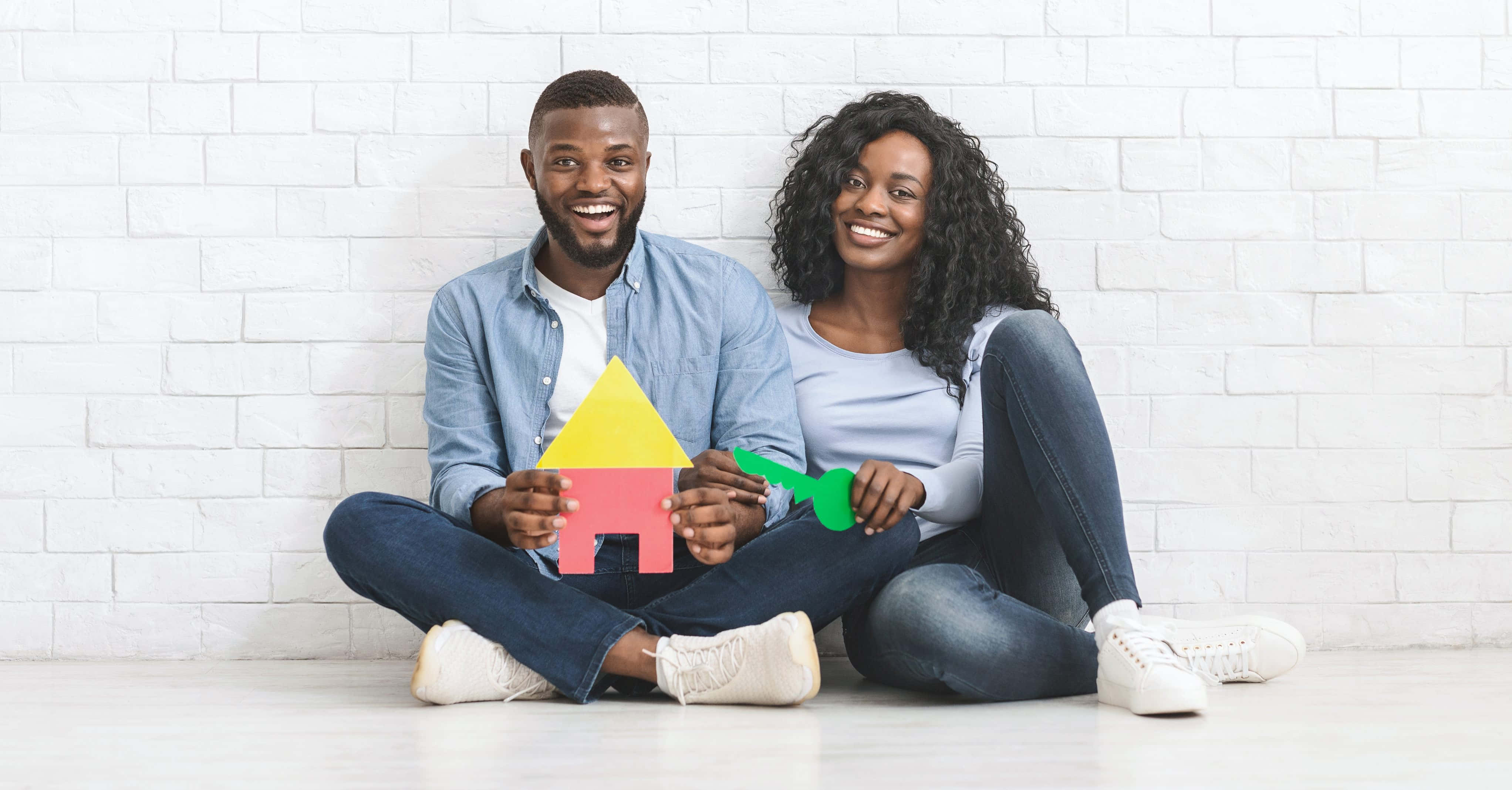 Black Couple 桌布 壁紙 — 一對微笑的情侶盤腿坐在白色地板上，對著白色磚牆手持色彩繽紛的紙屋和鑰匙剪紙。