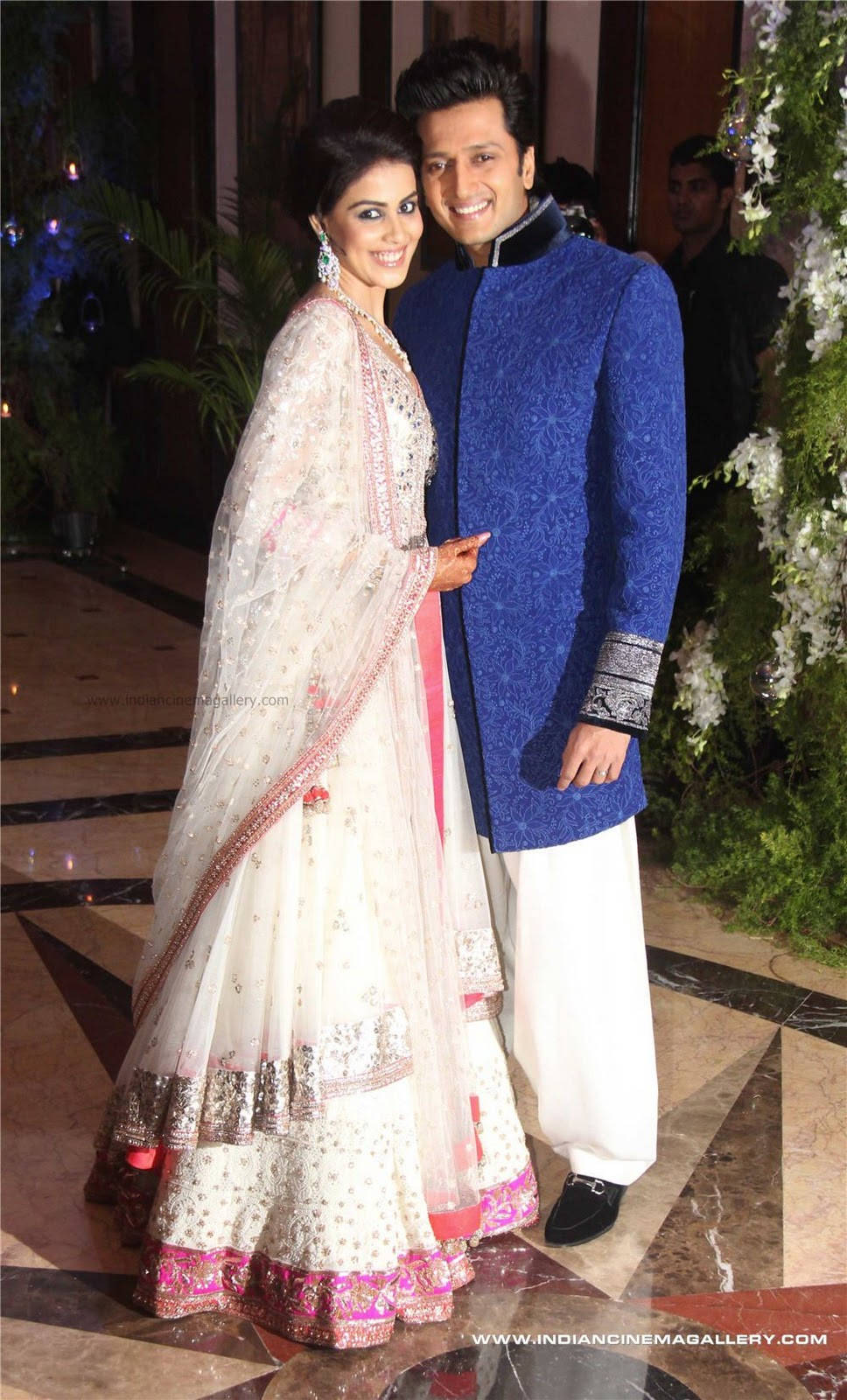 riteish deshmukh 桌布 壁紙 — 一對微笑的情侶穿著傳統印度服飾，在一個優雅的室內活動中拍照，場地有花卉裝飾和大理石地板。