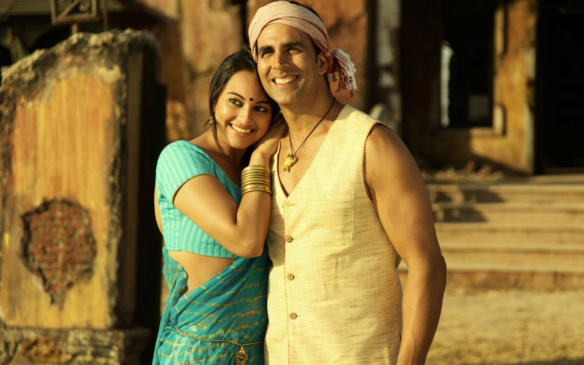 Akshay Kumar 桌布 壁紙 — 一對微笑的情侶穿著傳統印度服飾，在陽光明媚的質樸石結構前溫暖地擁抱。