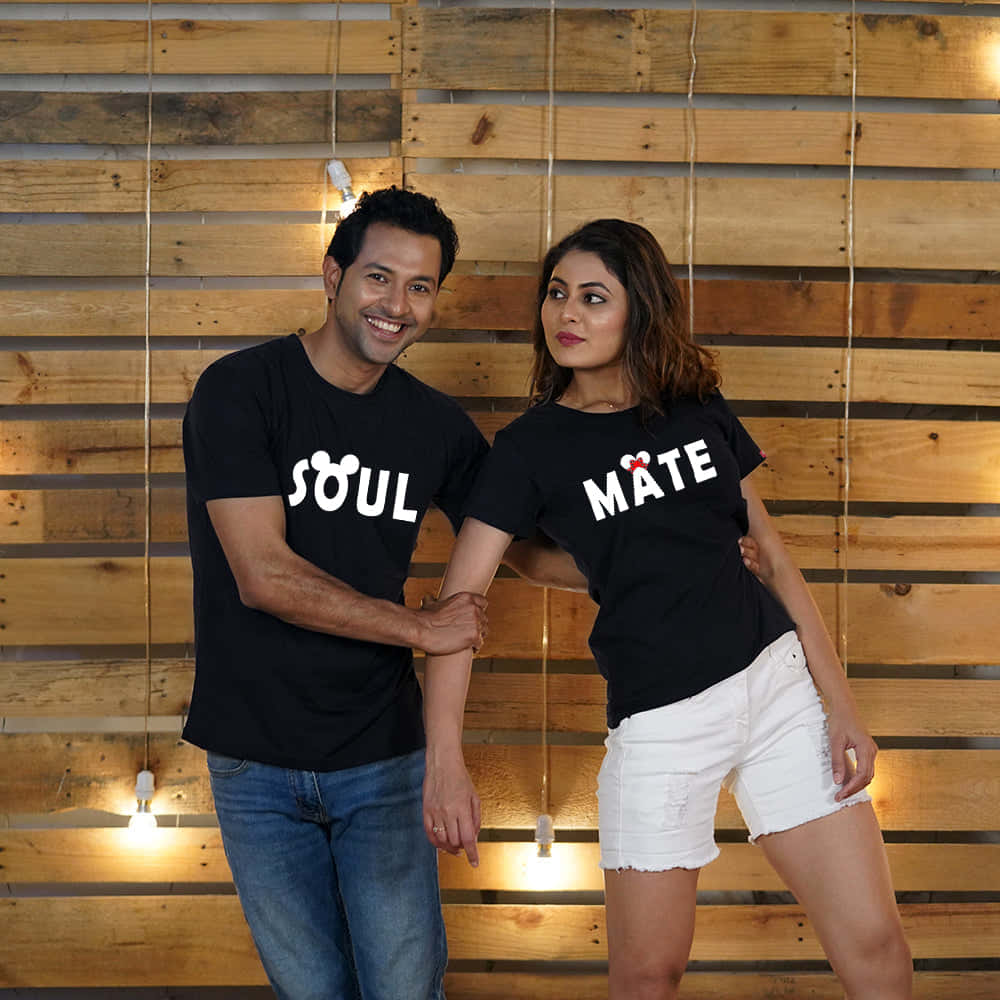 Romance 桌布 壁紙 — 一對微笑的情侶穿著印有「SOUL」和「MATE」字樣的黑色情侶T恤，站在溫暖的木牆前，牆上掛著燈串。