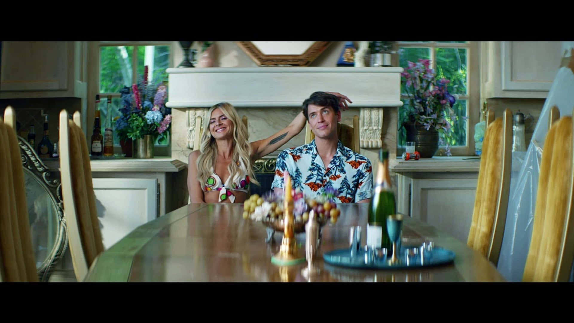 eliza coupe 桌布 壁紙 — 一對微笑的情侶穿著熱帶襯衫，坐在陽光充足的廚房裡，餐桌上擺放著鮮花和酒瓶。