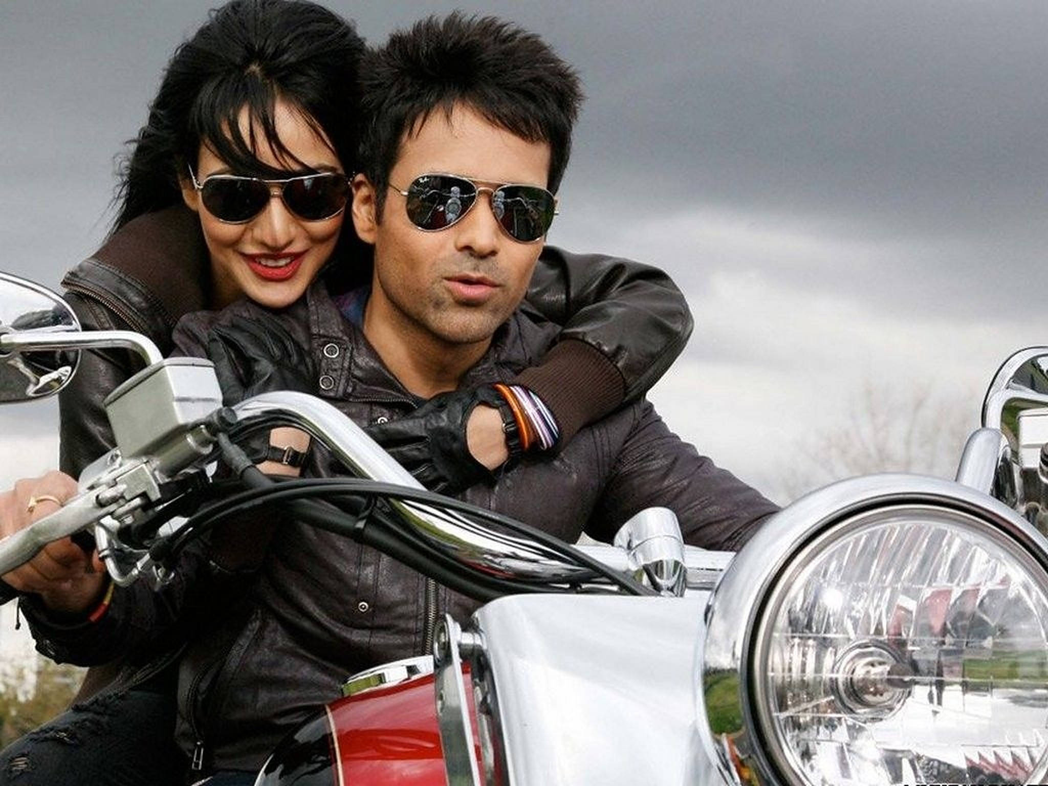 Emraan Hashmi 桌布 壁紙 — 一對微笑的情侶穿著皮夾克和太陽眼鏡，在多雲的天空下騎著一輛鍍鉻摩托車。