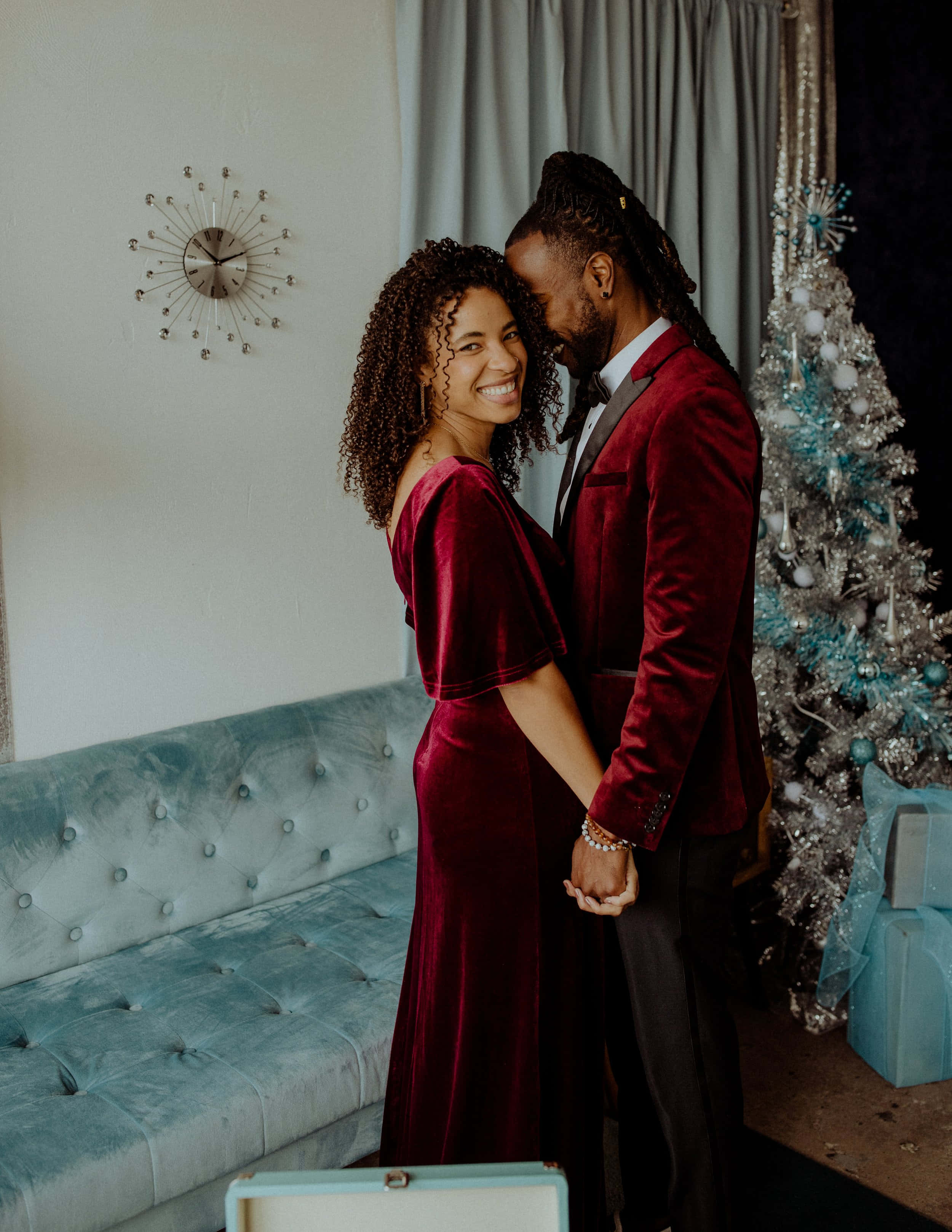 Couples Christmas 桌布 壁紙 — 一對微笑的情侶穿著相配的酒紅色天鵝絨服裝，在銀色聖誕樹和藍綠色沙發旁溫暖地擁抱。
