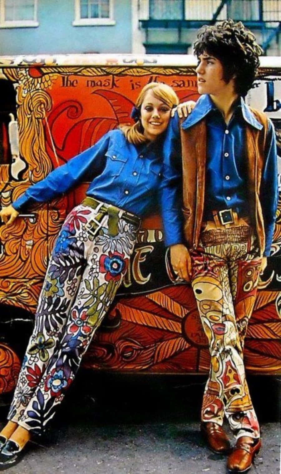 70年代動圖 桌布 壁紙 — 一對微笑的情侶穿著迷幻的1970年代服裝，在一輛塗有鮮豔漩渦圖案和文字的貨車旁擺姿勢。