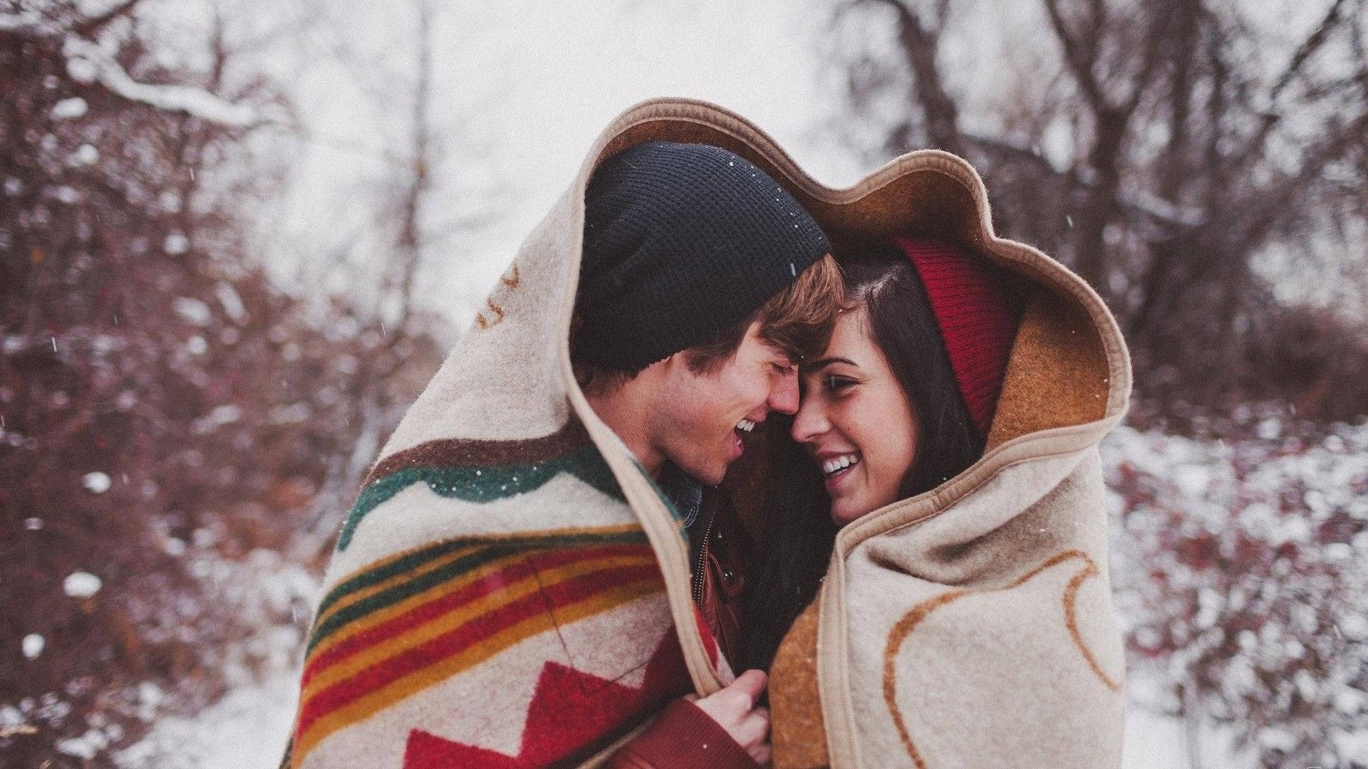 Love Story 桌布 壁紙 — 一對微笑的情侶裹著彩色毯子，在飄雪和光禿的冬日樹木間分享溫柔時刻。