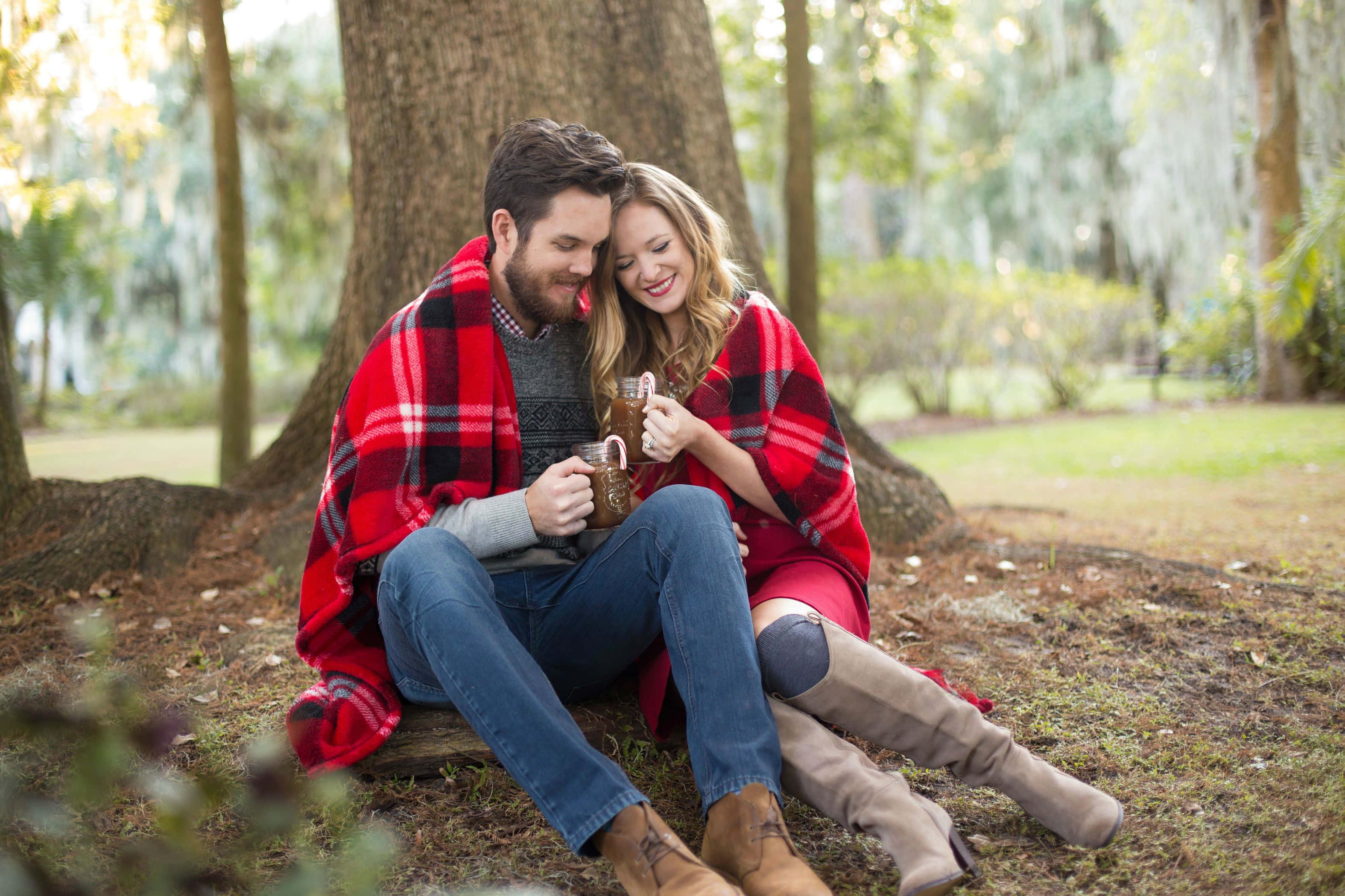 Christmas Couple 桌布 壁紙 — 一對微笑的情侶裹著紅色格子毯，坐在森林的樹根上，手持杯子。