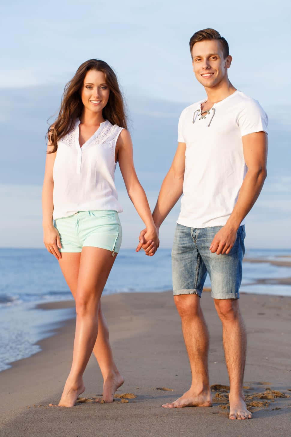 Holding Hands 桌布 壁紙 — 一對微笑的情侶赤腳在沙灘上牽手，身後有海浪和柔和天空。