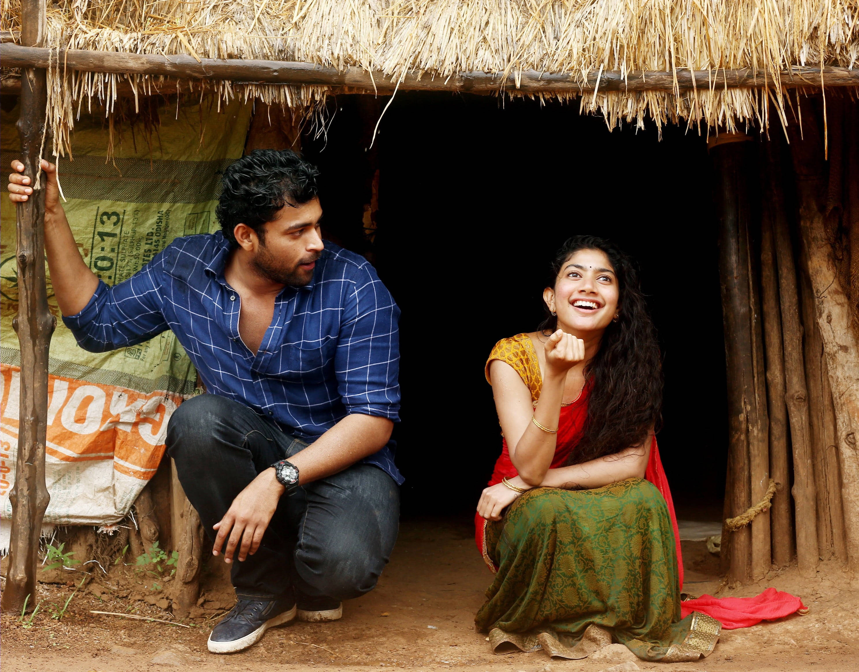 Sai Pallavi 桌布 壁紙 — 一對微笑的情侶蹲在鄉村茅草屋外，帶有溫暖的大地色調和自然質感。