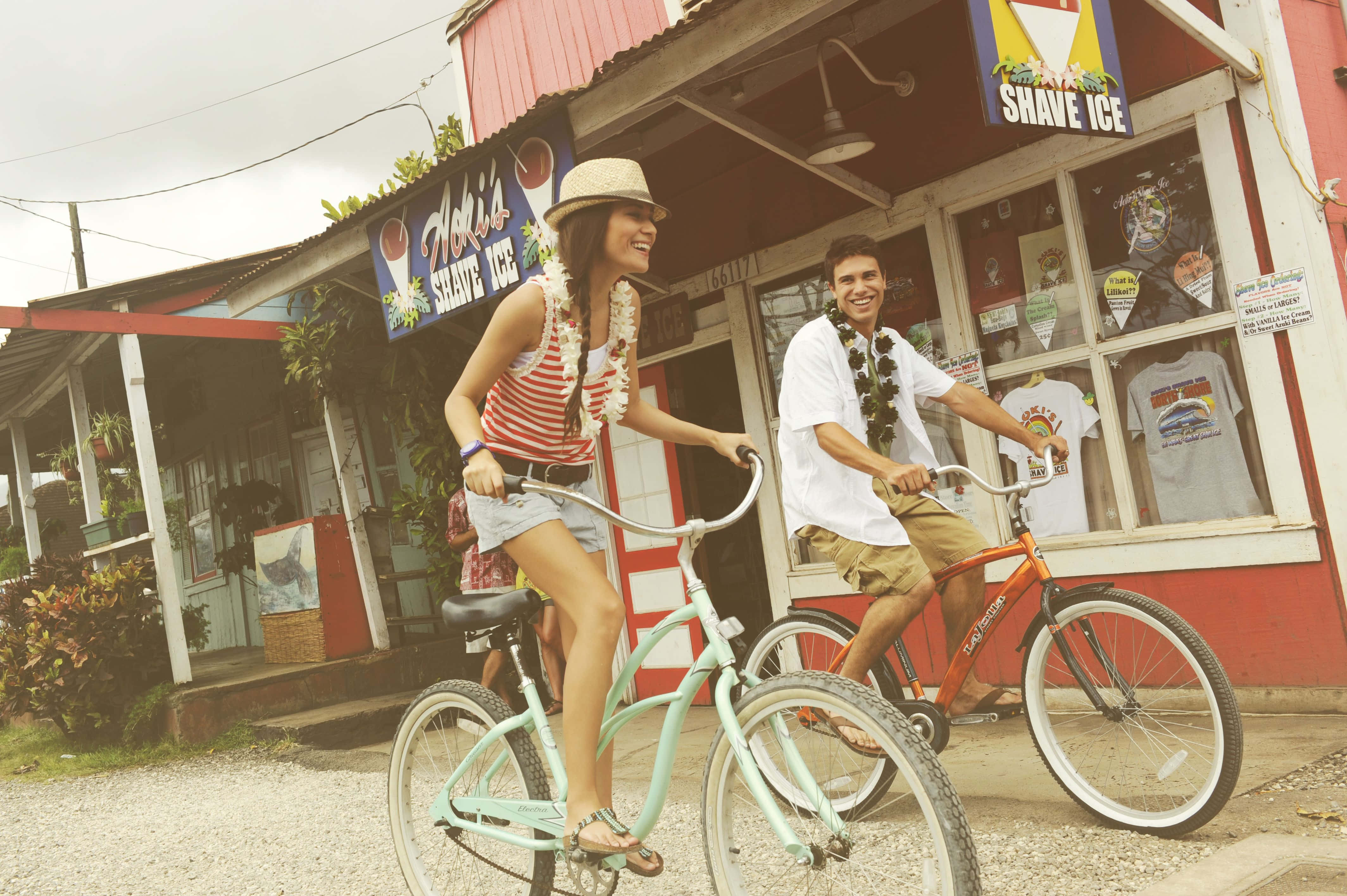 Beach Cruiser 桌布 壁紙 — 一對微笑的情侶騎腳踏車經過陽光明媚的熱帶街道上一間色彩繽紛的刨冰店。