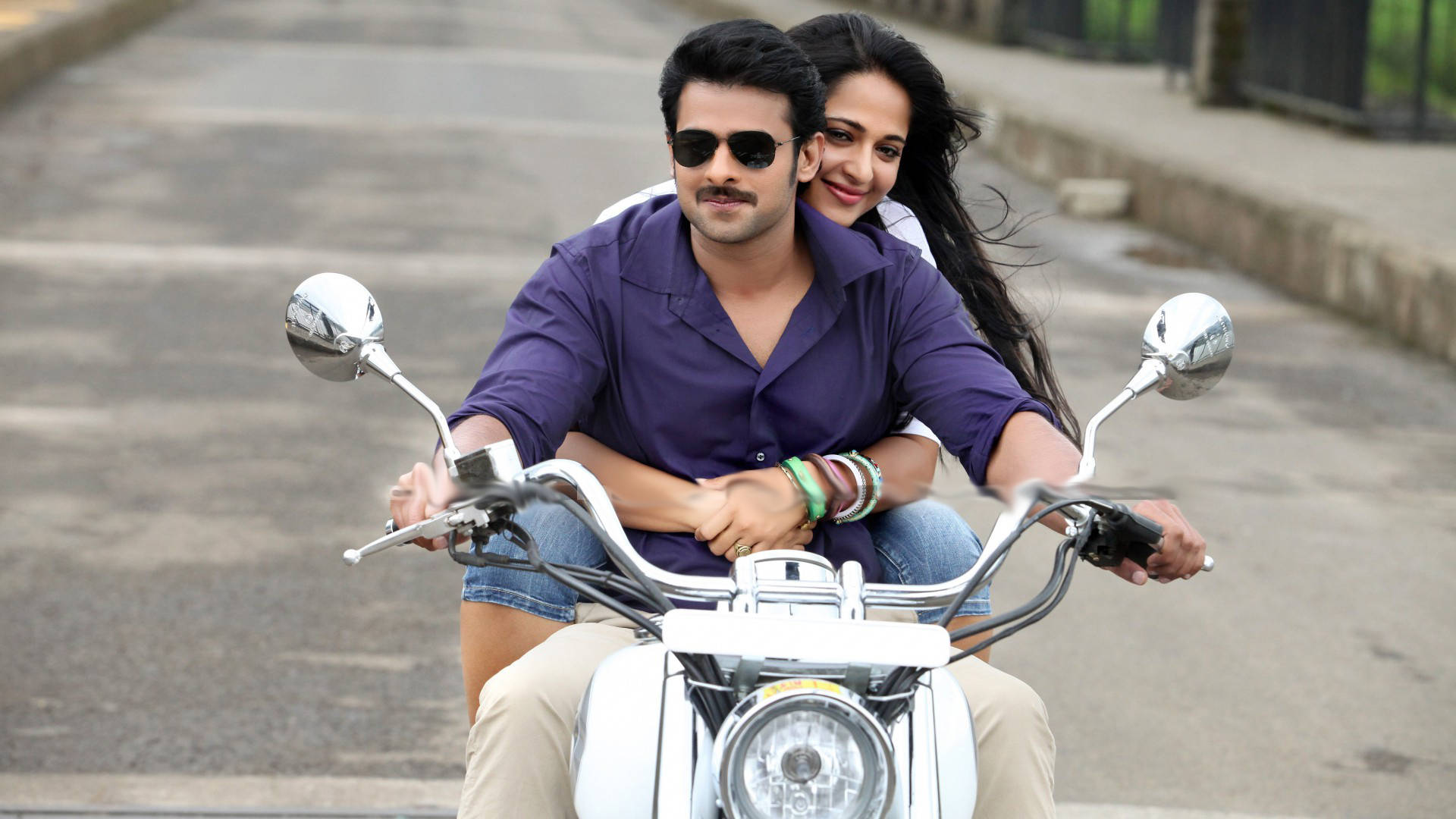 Prabhas HD 桌布 壁紙 — 一對微笑的情侶騎著白色機車行駛在鋪砌的街道上，背景有綠化。