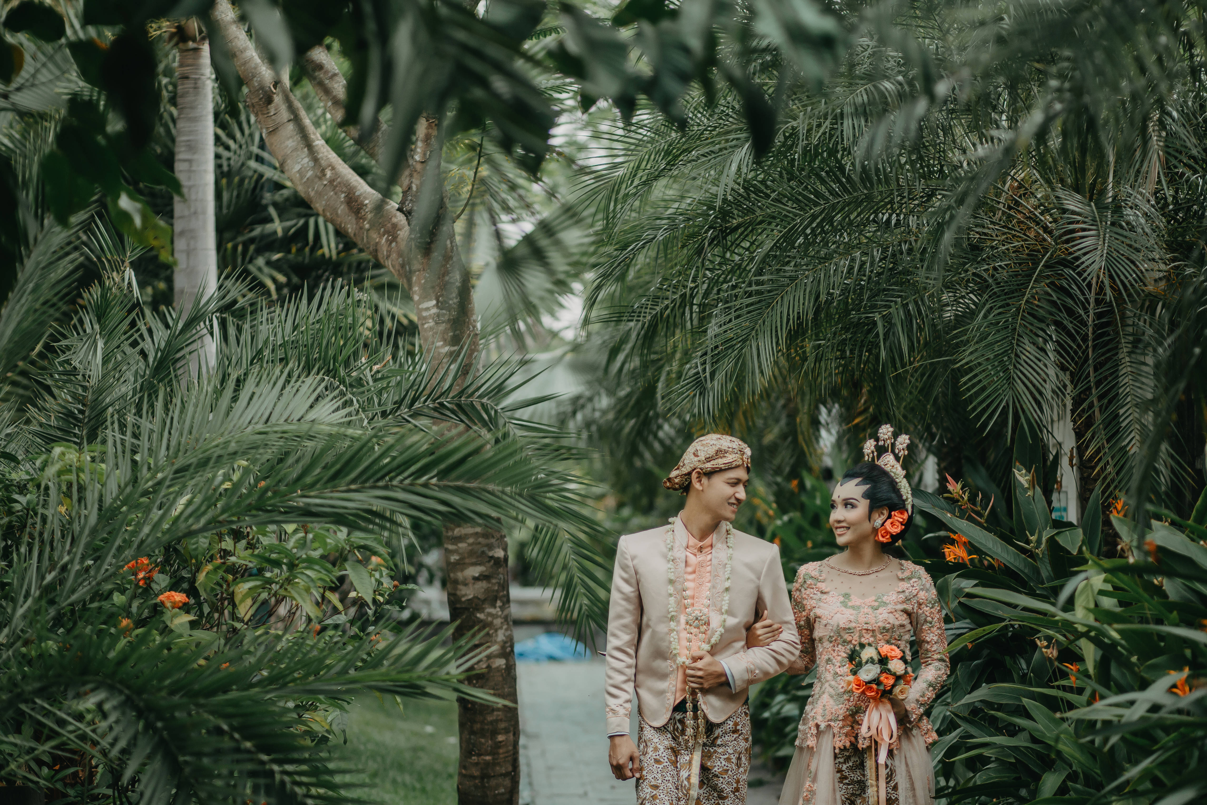 Groom 桌布 壁紙 — 一對微笑的新人穿著傳統峇里島婚禮服飾，手牽手走過棕櫚葉環繞的茂密熱帶花園。