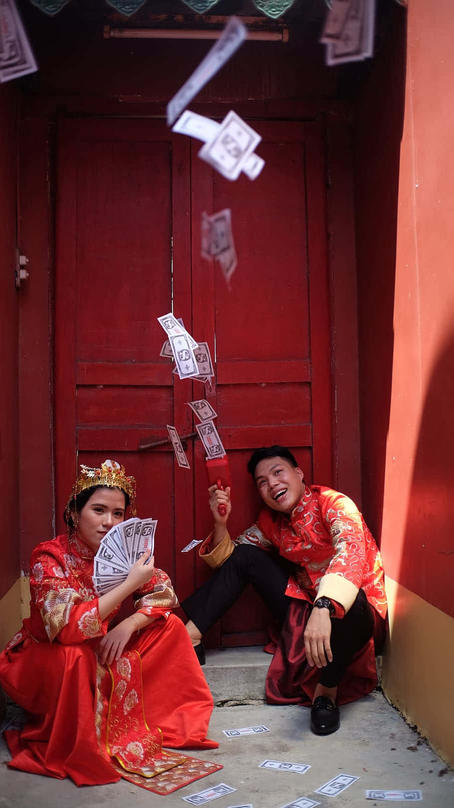 金錢 桌布 壁紙 — 一對微笑的新人穿著傳統紅色婚禮服飾，坐在紅色門前，慶祝時金錢在他們周圍飛舞。