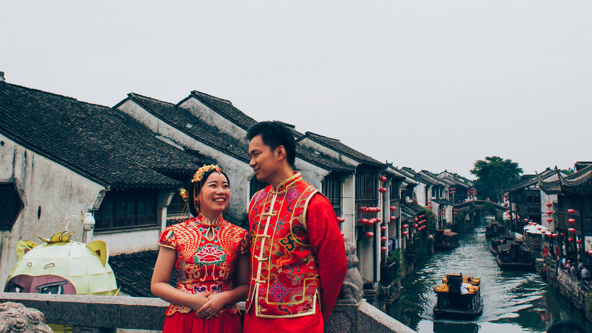 Suzhou 桌布 壁紙 — 一對微笑的新人穿著鮮豔的紅色傳統婚禮服飾，站在一座石橋上，俯瞰著兩旁歷史悠久的瓦頂房屋和漂浮船隻的運河。