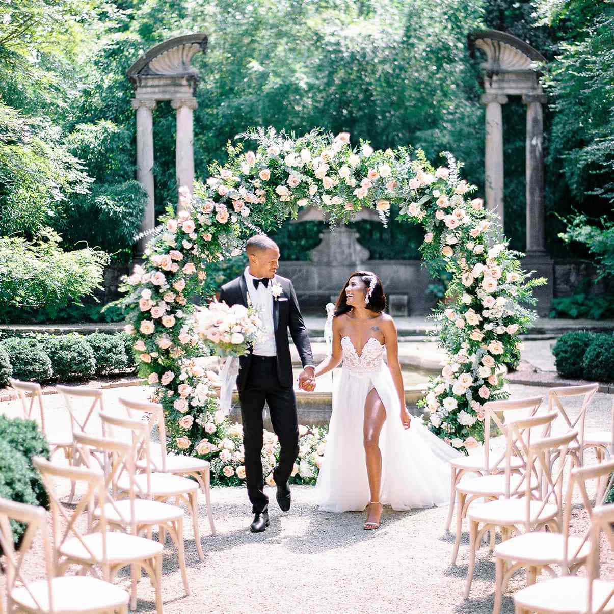 Garden Wedding 桌布 壁紙 — 一對微笑的新娘和新郎在陽光普照的花園婚禮中，手牽手走在茂盛的花拱門下。