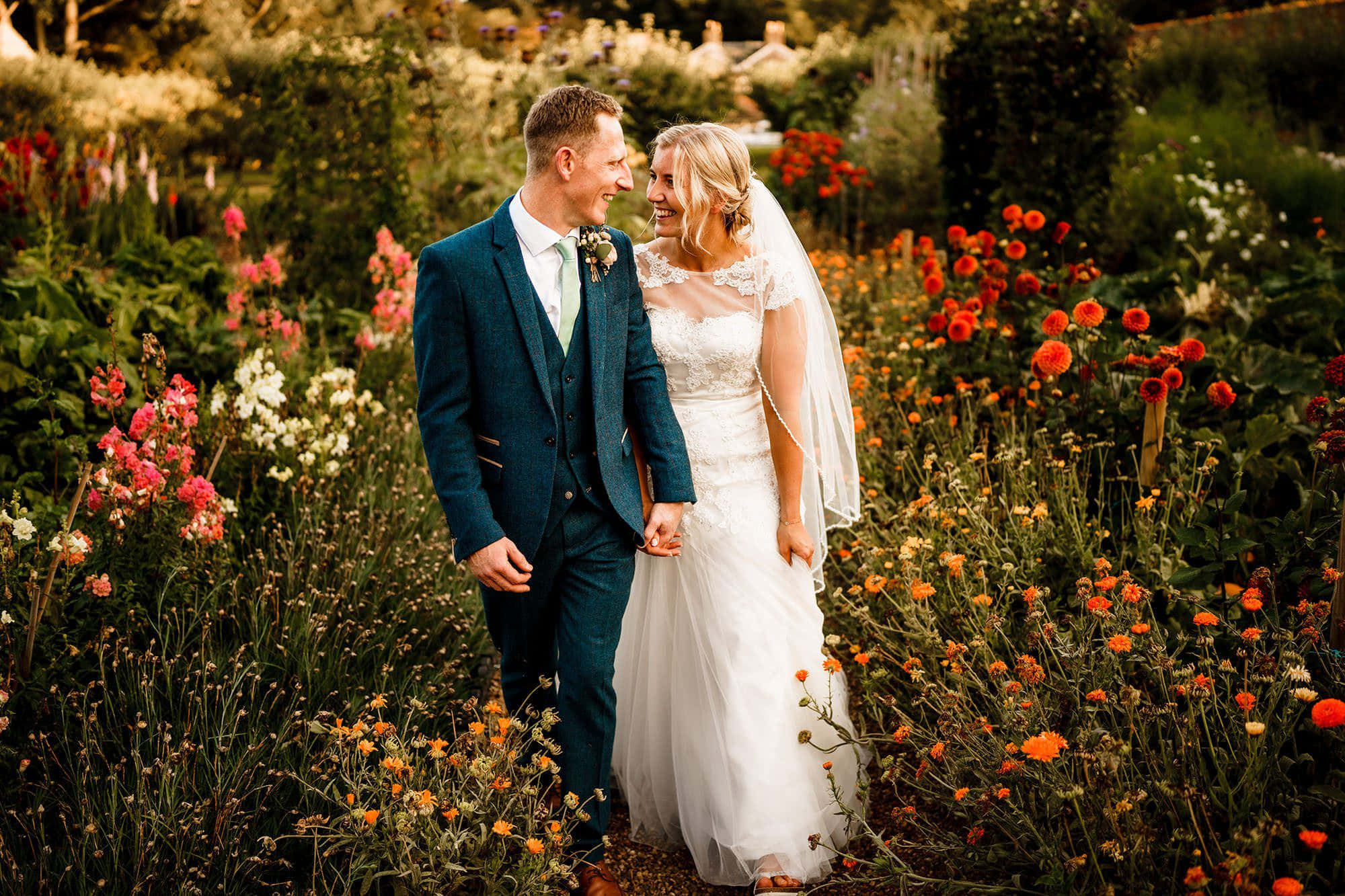 Garden Wedding 桌布 壁紙 — 一對微笑的新郎新娘手牽手，在陽光普照、鮮花盛開的花園中凝視對方。