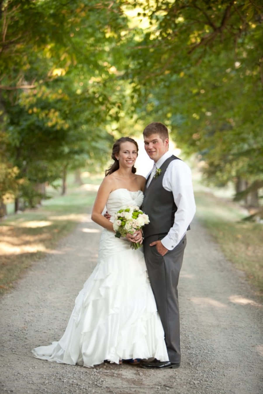 Wedding Couple 桌布 壁紙 — 一對微笑的新郎新娘站在陽光普照、樹木成蔭的碎石路上，手持花束。