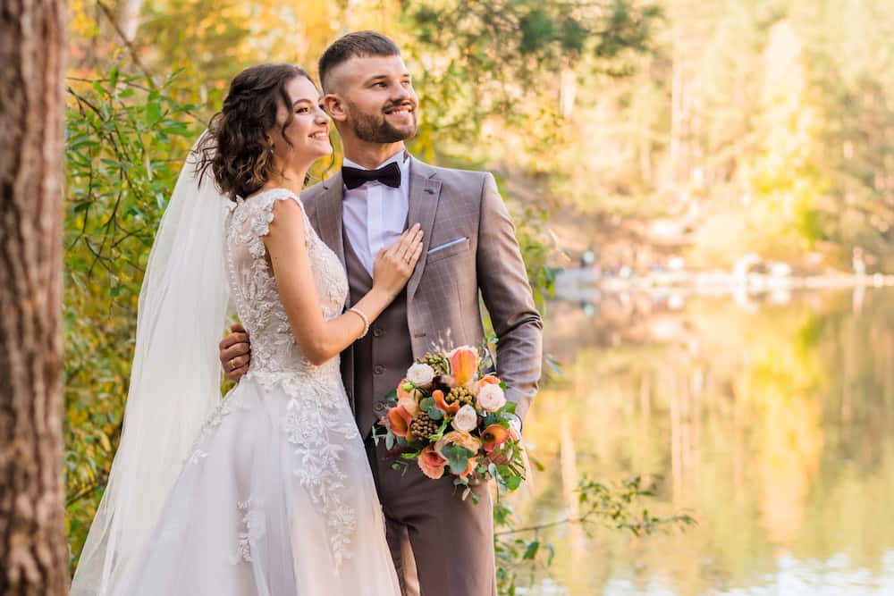 Groom And Bride 桌布 壁紙 — 一對微笑的新郎新娘站在陽光照耀的湖邊，周圍是秋天的樹木，手持色彩繽紛的花束。