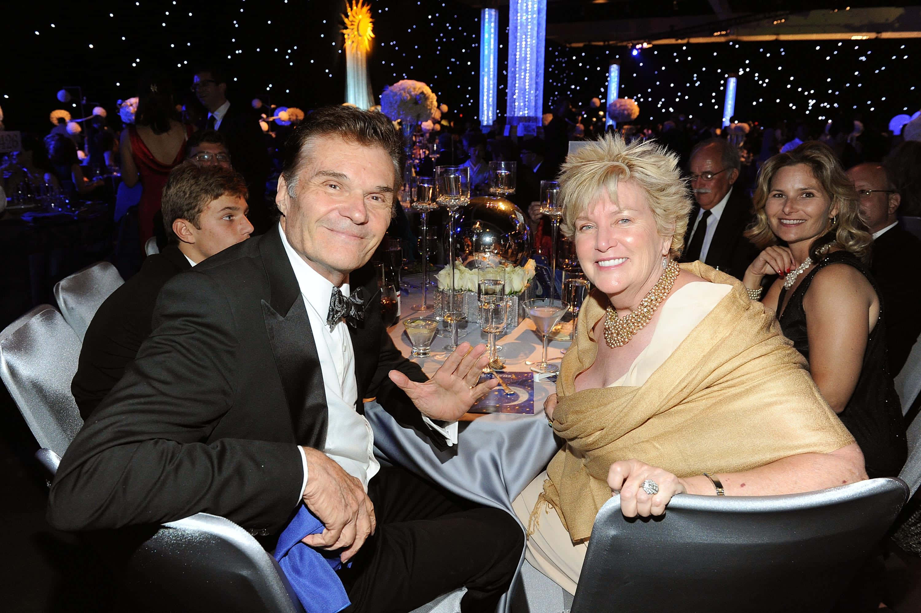 Fred Willard 桌布 壁紙 — 一對微笑的男女穿著正裝坐在閃閃發光的晚宴桌旁，周圍是賓客和閃爍的燈光。