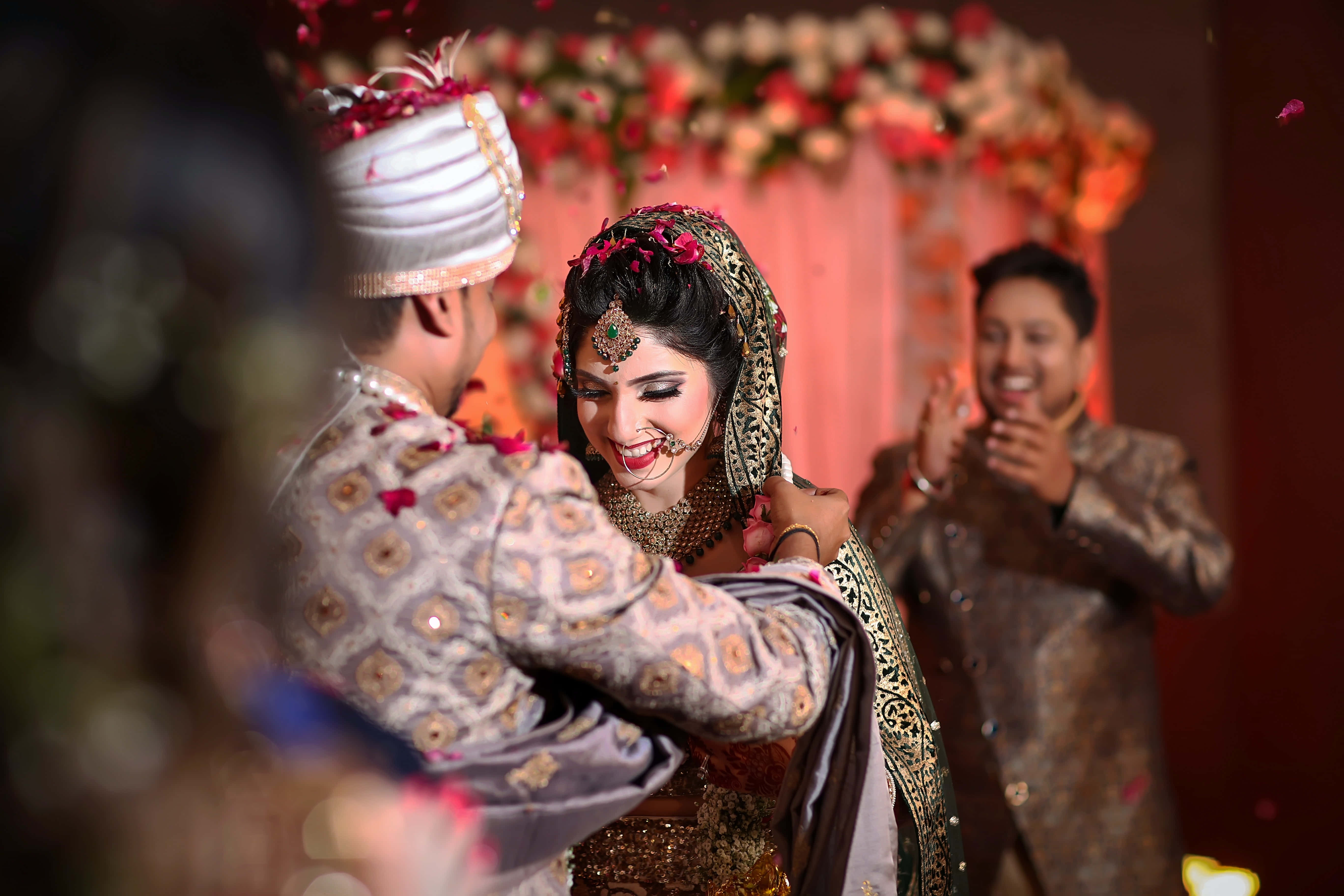 Wedding 桌布 壁紙 — 一對快樂的印度新娘和新郎在花拱門下擁抱，玫瑰花瓣如雨般灑落，在這場充滿活力的婚禮慶典中。