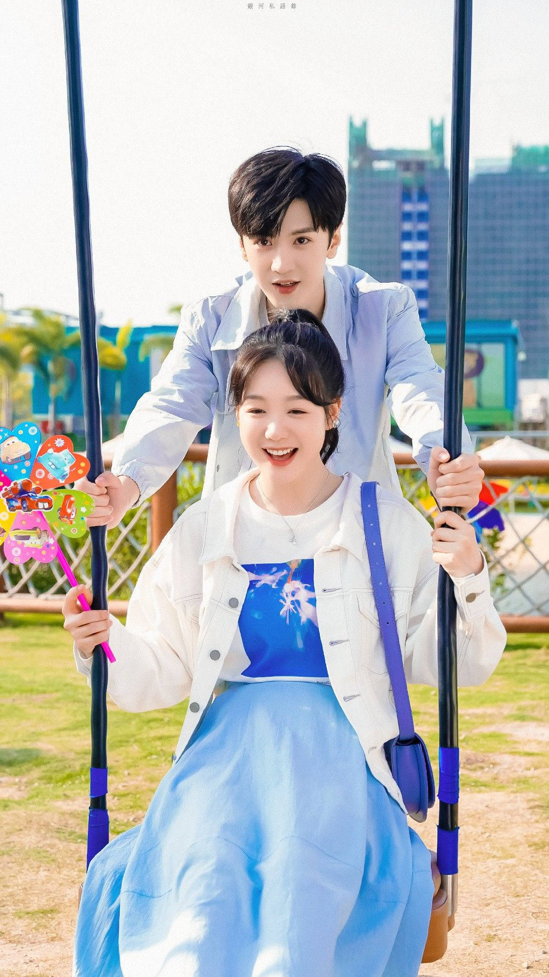 Couples 桌布 壁紙 — 一對快樂的年輕情侶在陽光明媚的城市公園中共享鞦韆，背景是現代建築。