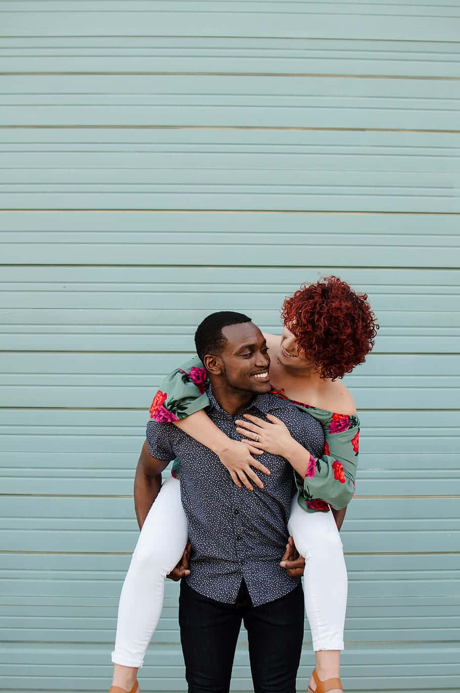 Interracial 桌布 壁紙 — 一對快樂的情侶在一個青綠色的波紋牆前，女方騎在男方背上，分享一個俏皮的吻。
