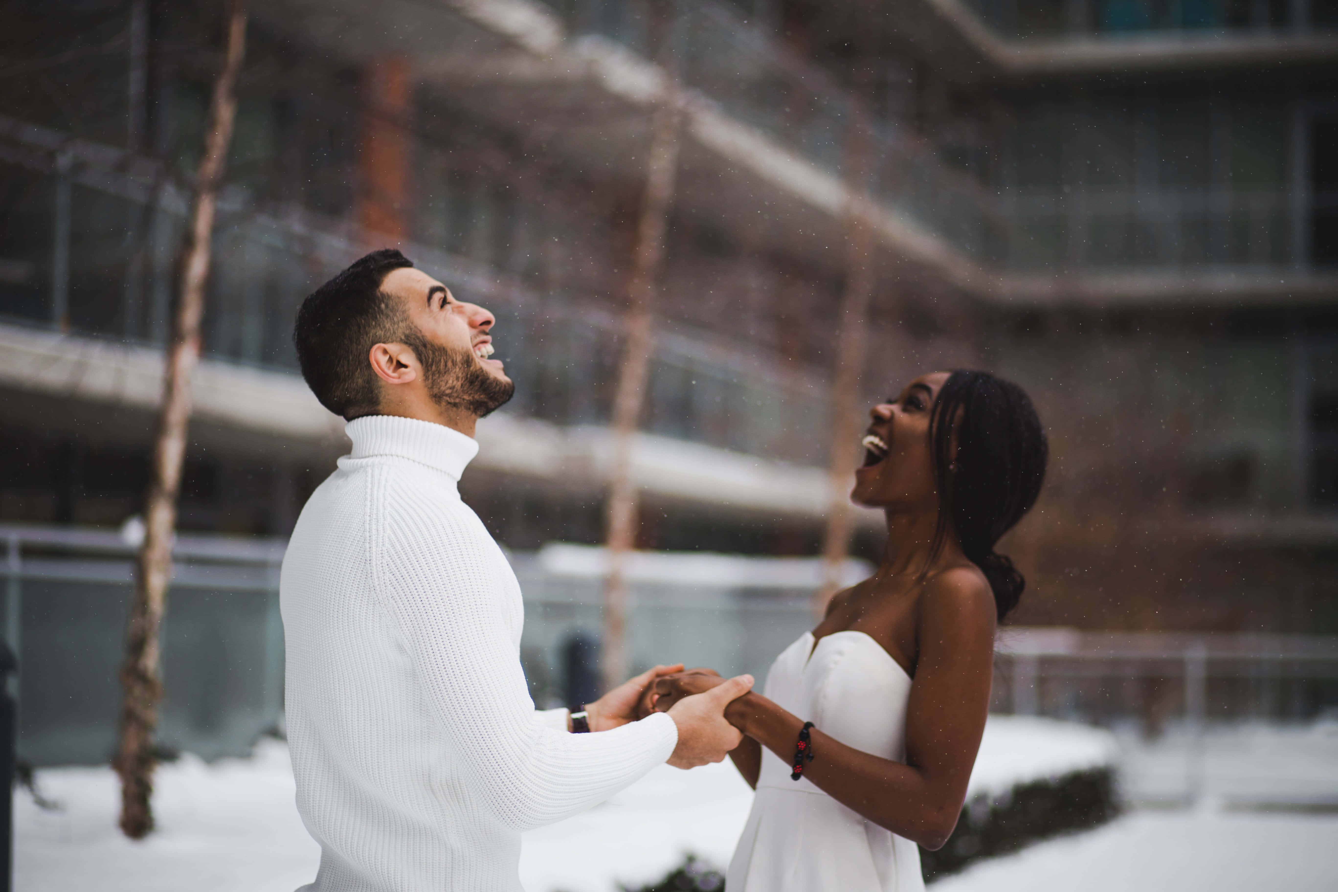 Interracial 桌布 壁紙 — 一對快樂的情侶在現代建築背景下，在飄落的雪中一起大笑。