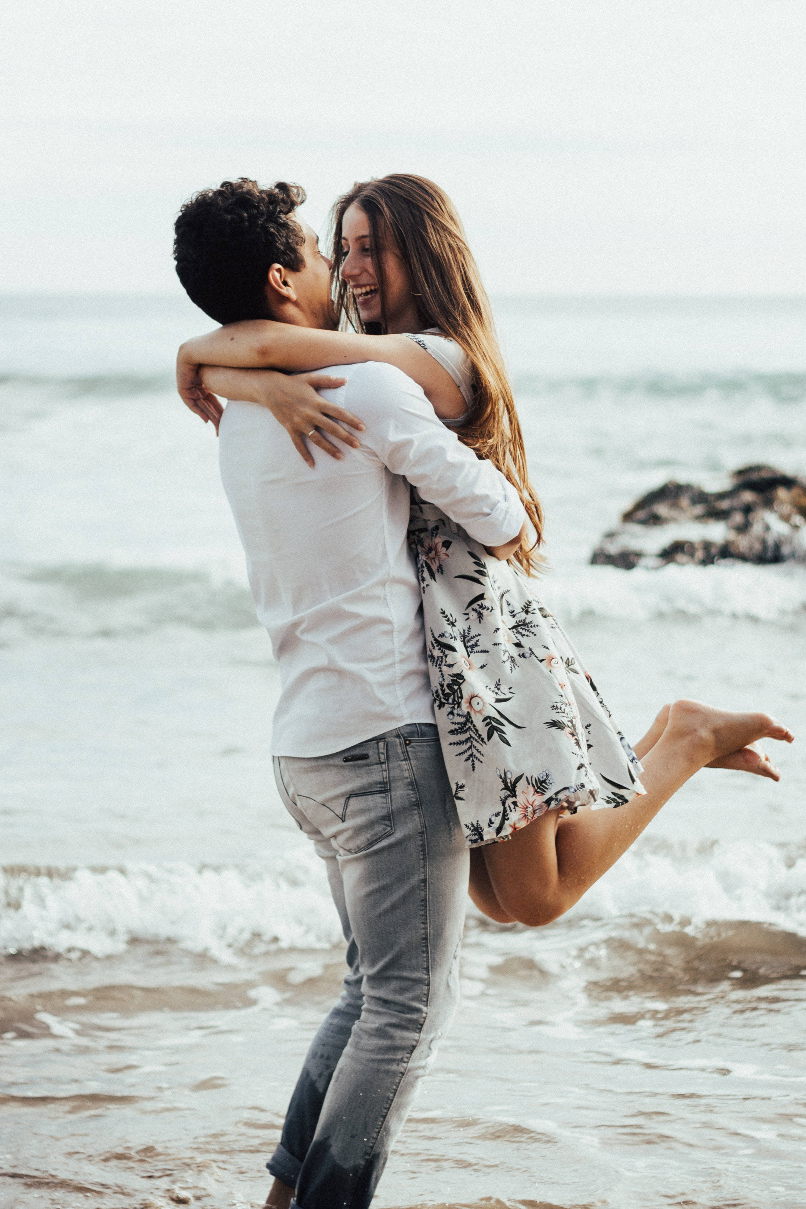 Couple Hugging 桌布 壁紙 — 一對快樂的情侶在陽光明媚的海灘上擁抱，背景是拍岸的浪花和遠處的岩石，天空柔和。