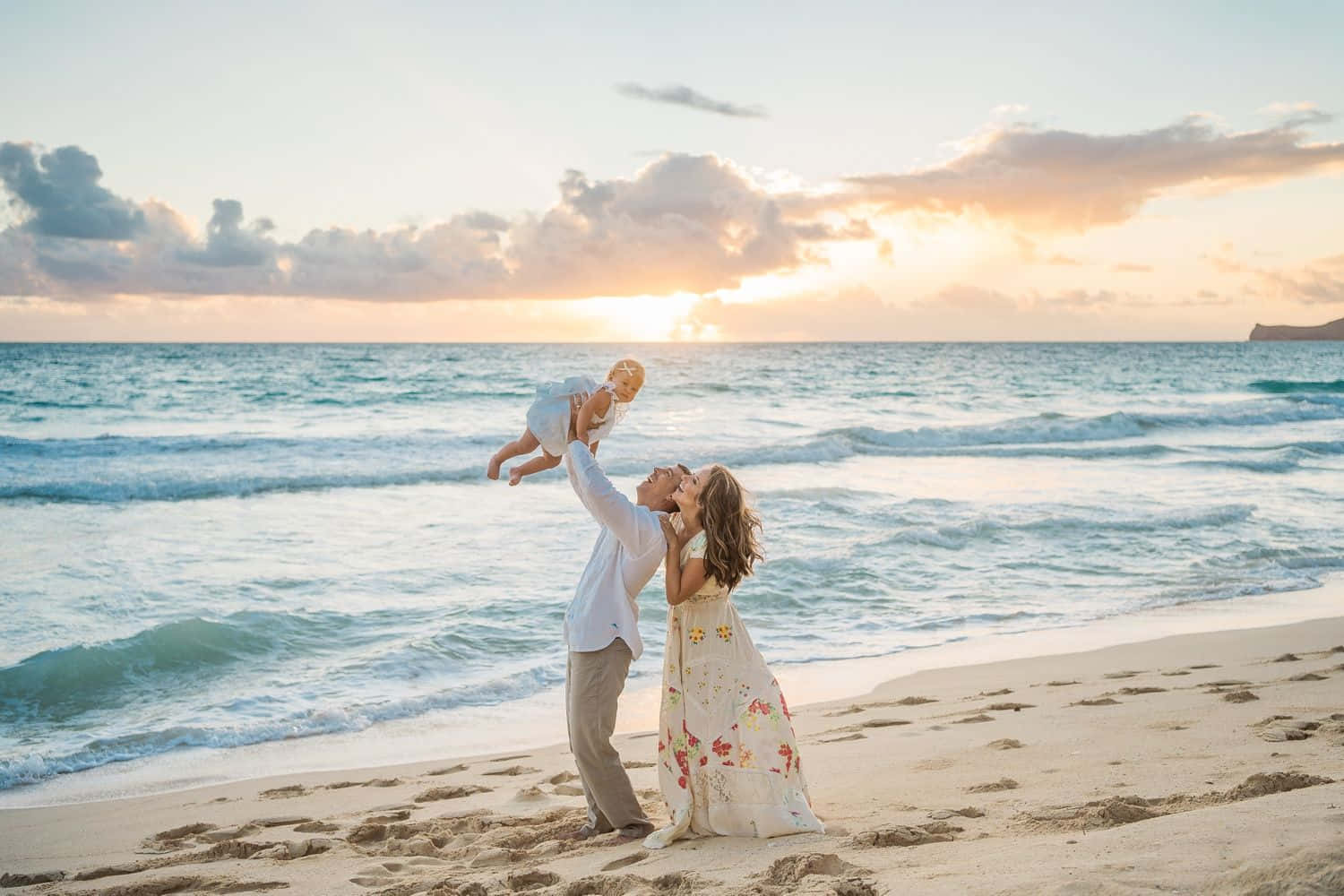 Family Beach 桌布 壁紙 — 一對快樂的情侶在陽光明媚的海灘上嬉戲地將嬰兒舉到空中，海浪拍打，夕陽將天空染成金色。