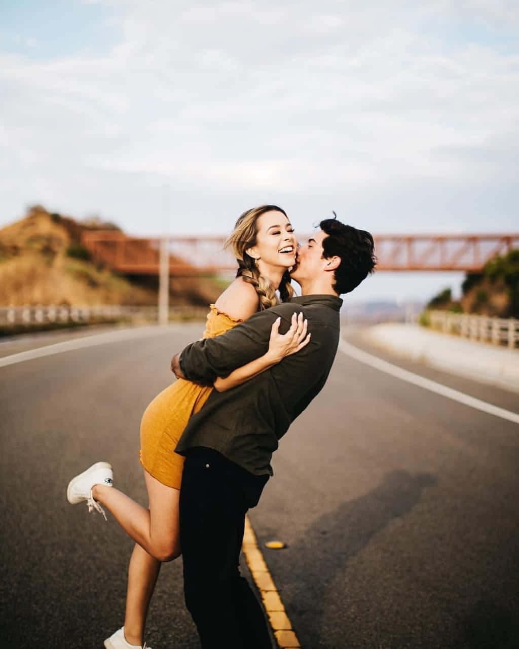 Cute Love 桌布 壁紙 — 一對快樂的情侶在陽光明媚的高速公路上分享一個俏皮的吻，背景是一座橋。