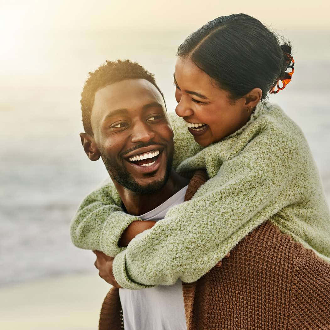 Romance 桌布 壁紙 — 一對快樂的情侶在黃金時段於海邊分享有趣的背人遊戲，溫暖的陽光突顯他們的笑聲。