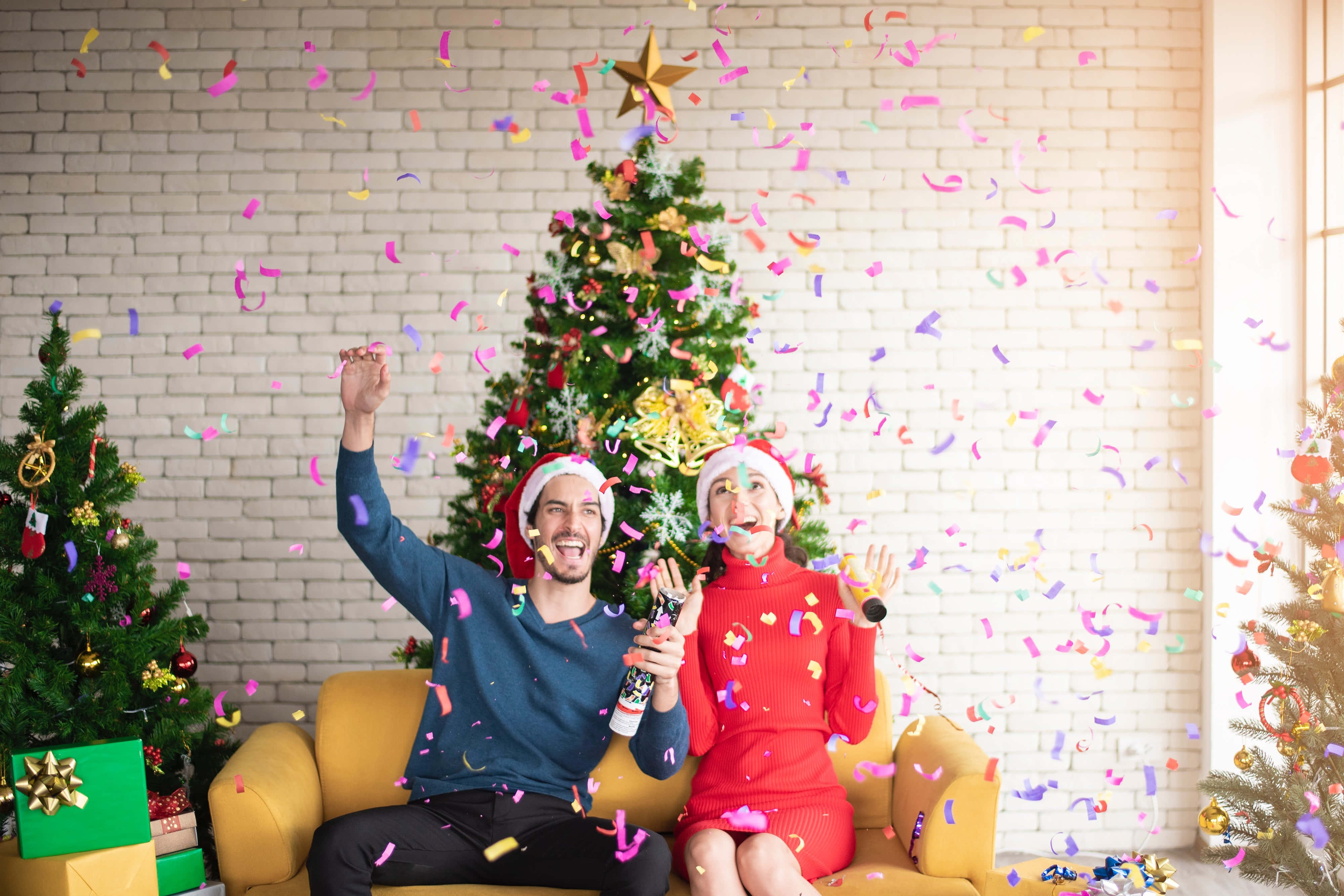 Couples Christmas 桌布 壁紙 — 一對快樂的情侶坐在黃色沙發上，旁邊有裝飾好的聖誕樹，周圍飄灑著五彩紙屑，慶祝聖誕節。