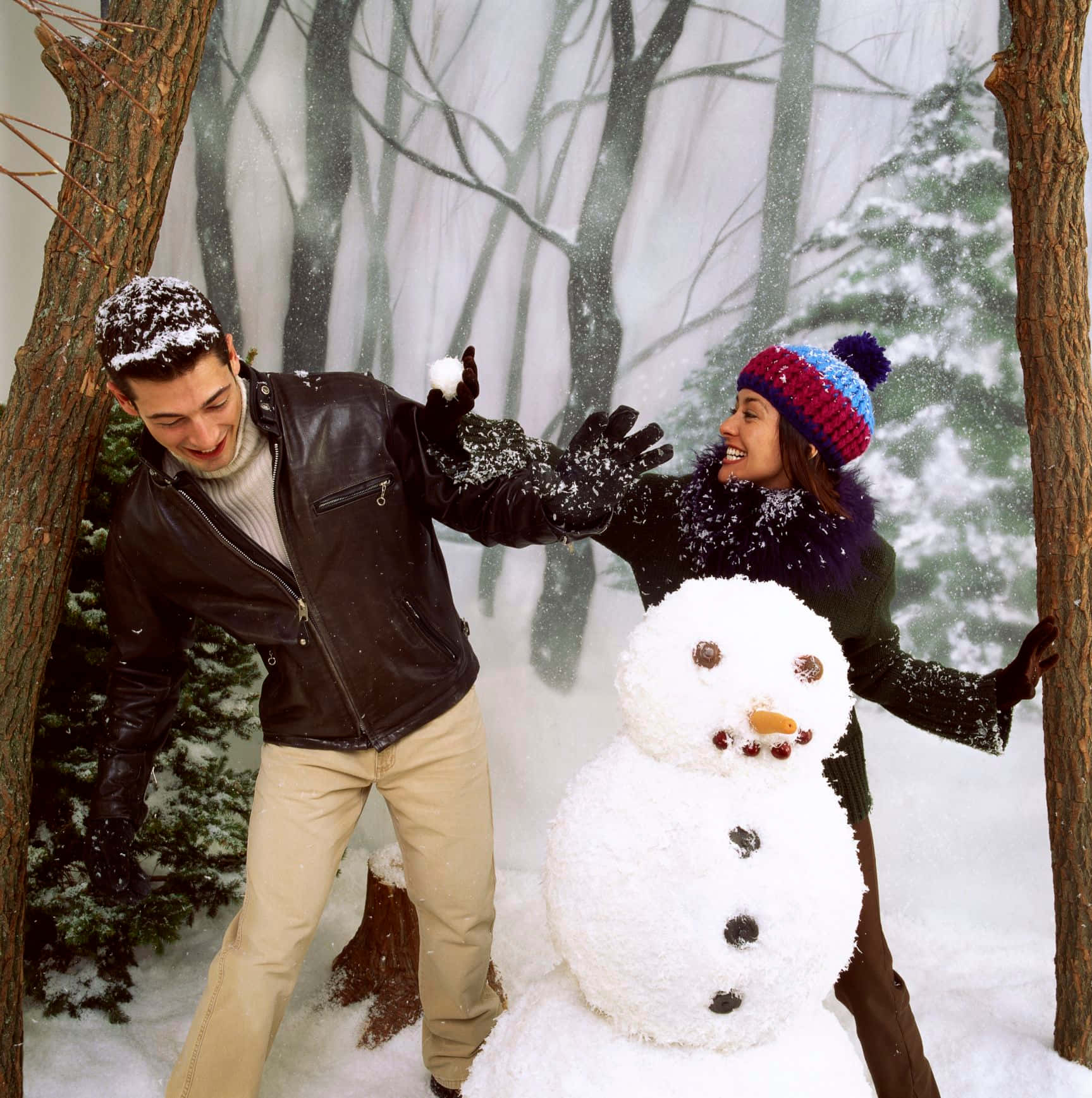 Couples Christmas 桌布 壁紙 — 一對快樂的情侶穿著冬季裝備，在飄雪中與雪人嬉戲於雪地森林。
