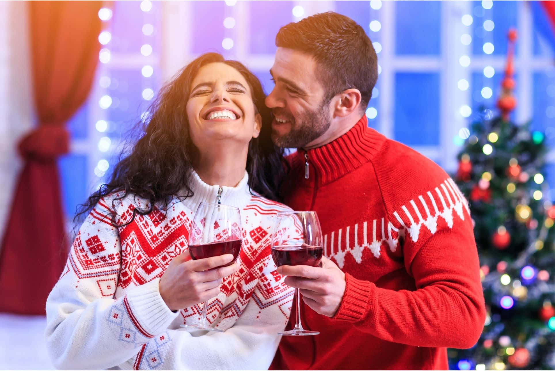 Couples Christmas 桌布 壁紙 — 一對快樂的情侶穿著節日毛衣，在裝飾好的聖誕樹附近拿著酒杯笑著。