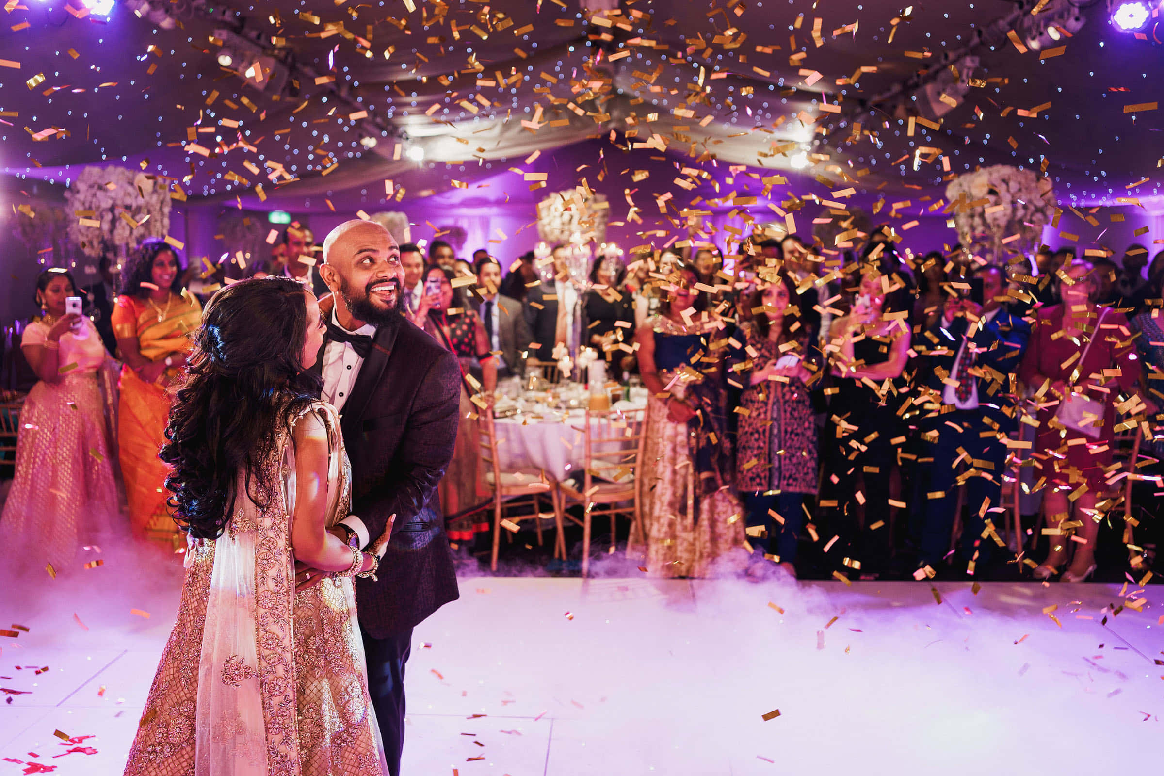 Bride And Groom 桌布 壁紙 — 一對快樂的新人在充滿活力的紫色燈光婚宴上，在飄落的金色紙屑下跳舞。