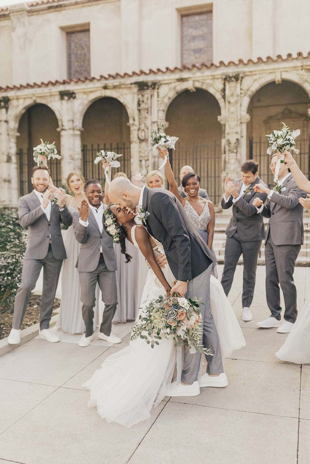 Bridal 桌布 壁紙 — 一對快樂的新人在陽光普照的庭院中分享一個俏皮的吻，賓客們歡呼並舉起花束。