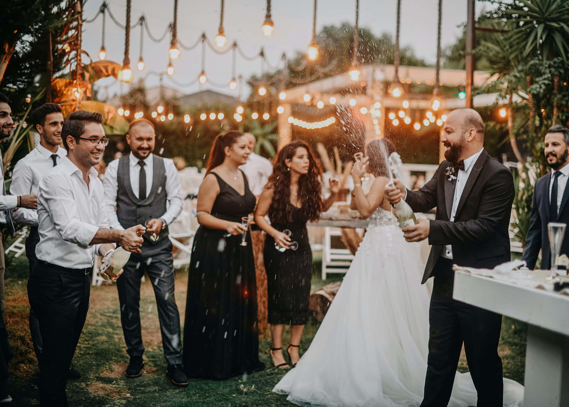 Garden Wedding 桌布 壁紙 — 一對快樂的新人在黃金時刻的茂密花園中，在串燈下用香檳慶祝。
