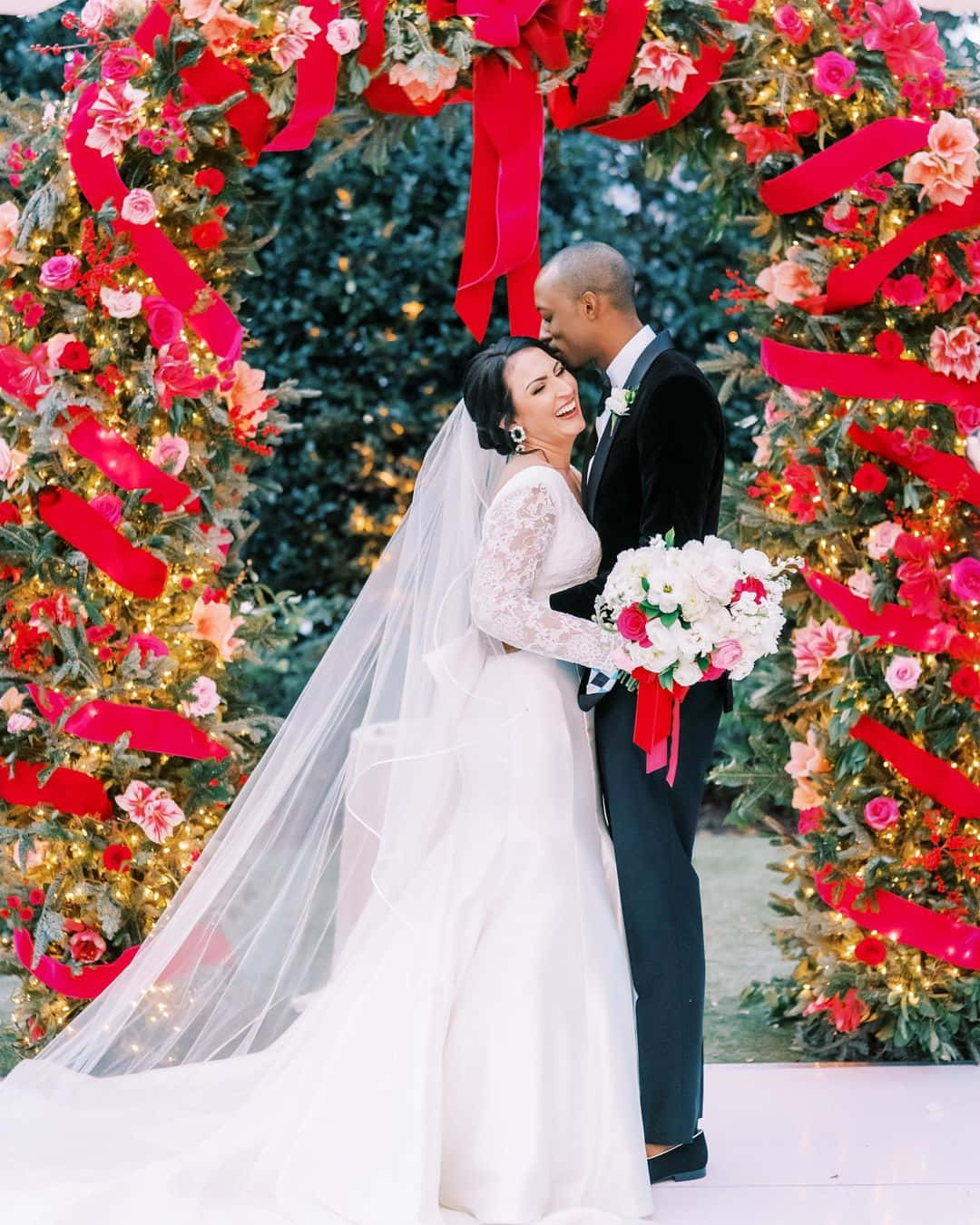 Bridal 桌布 壁紙 — 一對快樂的新娘和新郎在裝飾著閃爍燈光的節日紅色絲帶和鮮花拱門下擁抱。