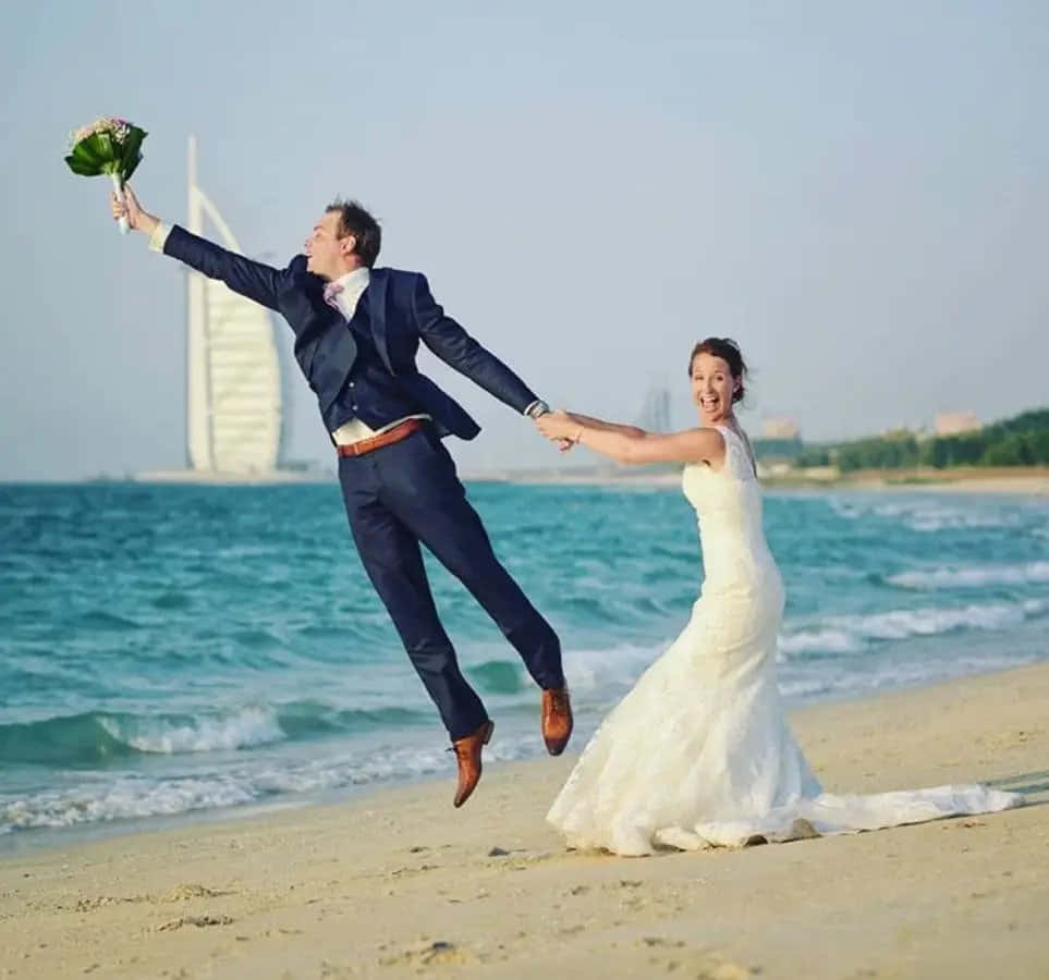 Funny Wedding 桌布 壁紙 — 一對快樂的新娘和新郎在陽光明媚的海灘上跳躍，背景是帆船酒店。