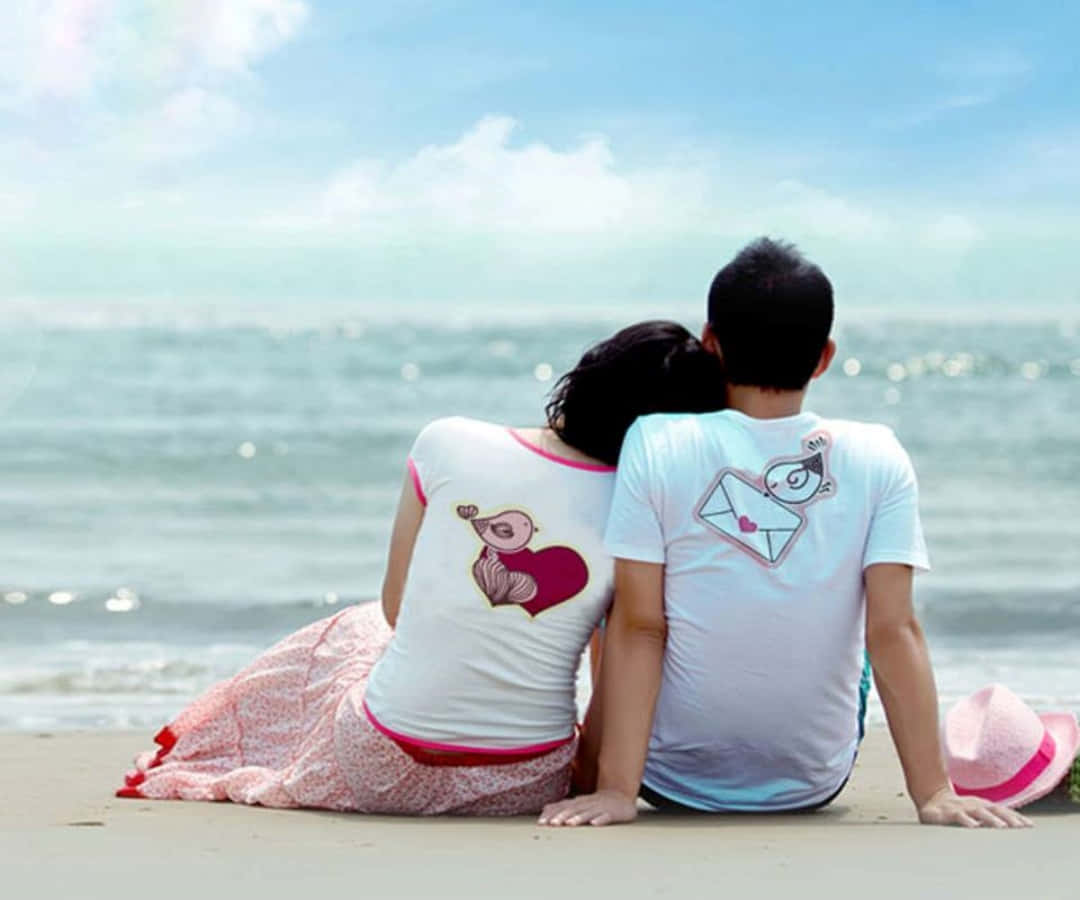 lover 桌布 壁紙 — 一對情侶並肩坐在陽光明媚的海灘上，面向海洋，穿著相配的可愛情侶主題圖案襯衫。