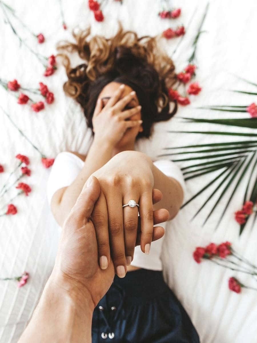 engagement 桌布 壁紙 — 一對情侶分享親密時刻，一隻手輕輕握住另一隻戴著訂婚戒指的手，周圍散落著紅花和棕櫚葉。