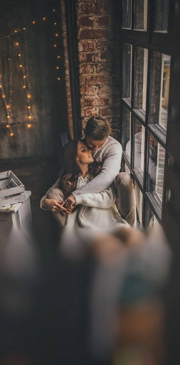 couple cuddle 桌布 壁紙 — 一對情侶在一個舒適的磚牆房間裡，在大窗戶旁溫柔地擁抱，房間裝飾著溫暖的燈串。