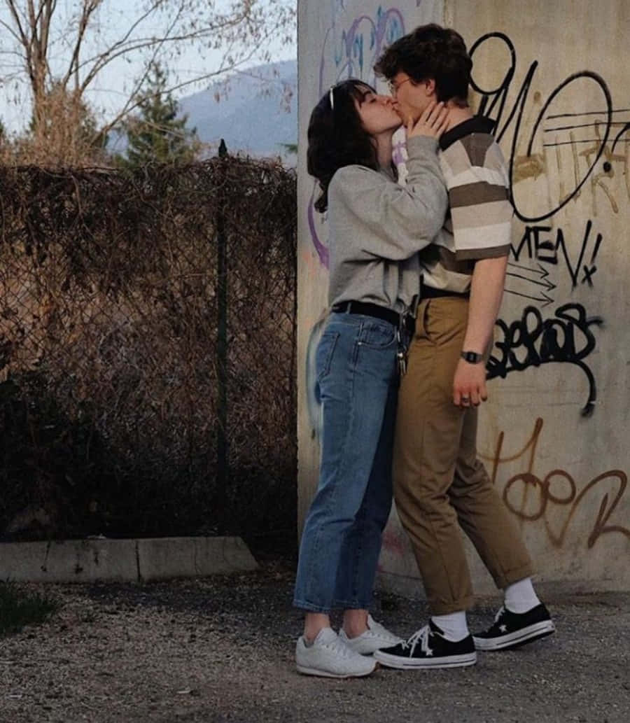 Aesthetic Couple 桌布 壁紙 — 一對情侶在佈滿塗鴉的牆前熱情接吻，背景有光禿的樹木和遠山，光線柔和。