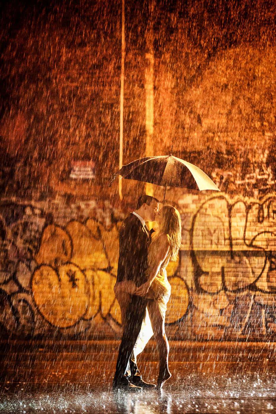 Couple In Rain 桌布 壁紙 — 一對情侶在傾盆大雨中於傘下接吻，背景是塗鴉牆，沐浴在溫暖的橙色燈光中。