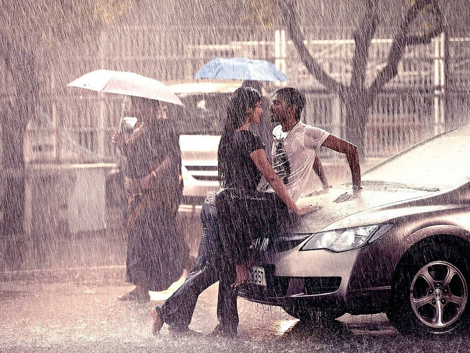 Couple 桌布 壁紙 — 一對情侶在傾盆大雨中靠著一輛銀色汽車分享親密時刻，附近另一個人撐著雨傘。