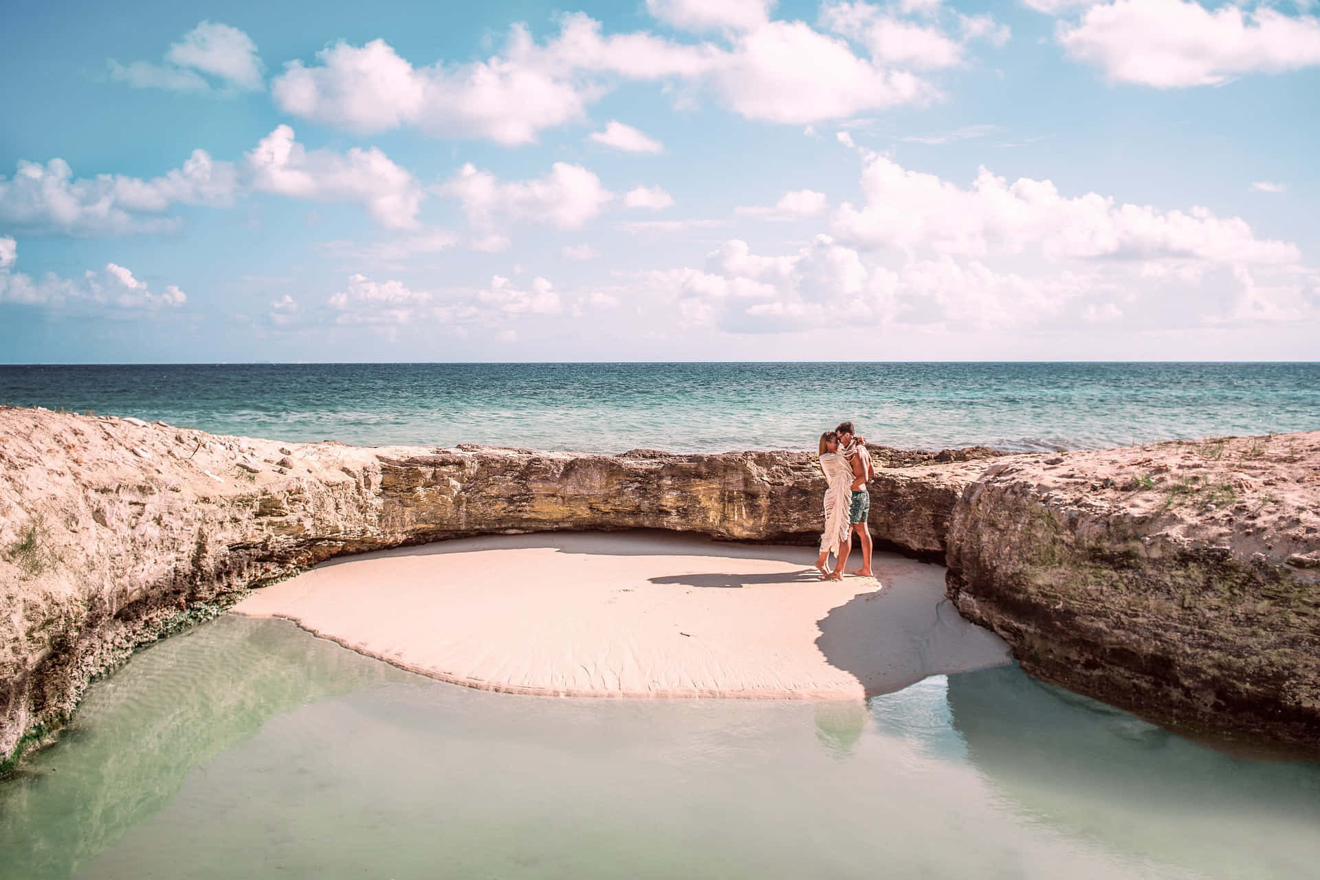 Cancun 桌布 壁紙 — 一對情侶在僻靜的沙灣中擁抱，四周是崎嶇的懸崖，頭頂是明亮的藍天和蓬鬆的雲朵。